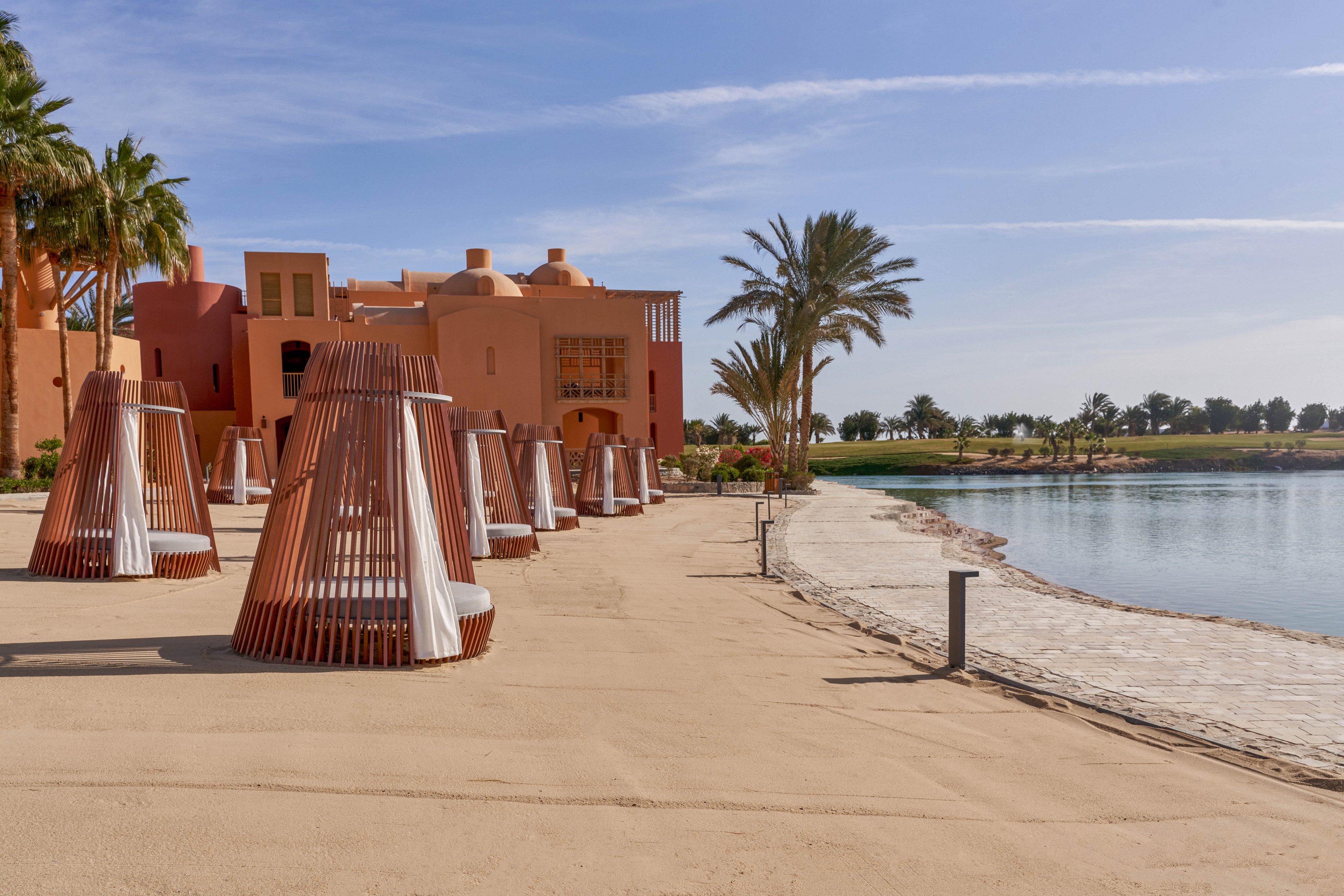 A serene beachfront resort featuring modern cabanas with wooden frames and white drapes. The setting includes sandy pathways, palm trees, and a tranquil water body under a clear blue sky. The architecture showcases warm terracotta tones, complementing the natural surroundings.