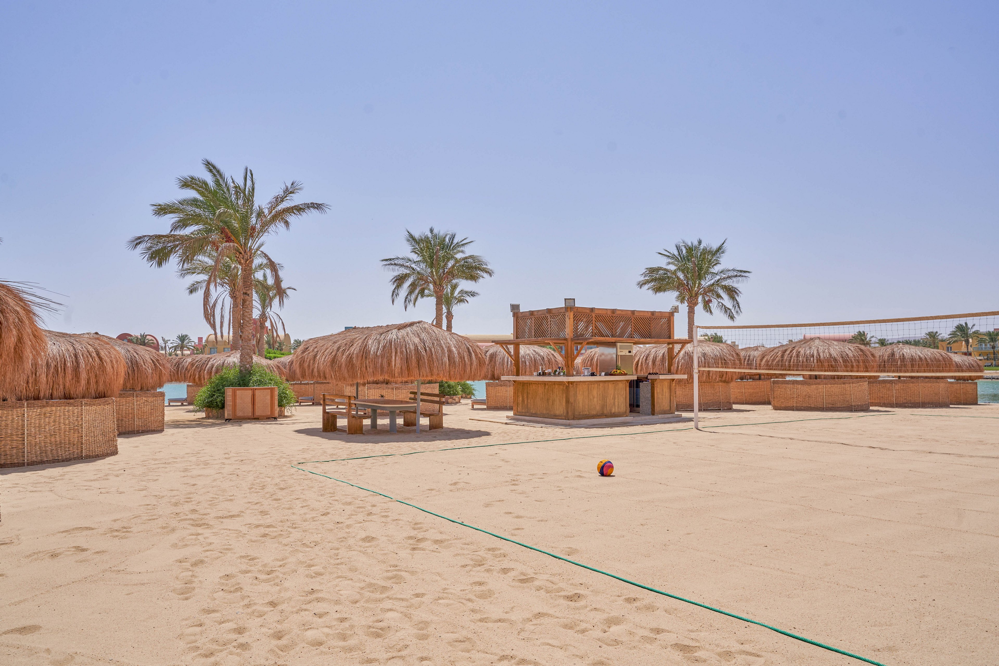 A sandy beach volleyball court surrounded by thatched-roof huts and tall palm trees under a clear blue sky. The setting exudes a tropical and relaxed vibe, with a volleyball placed on the sand. The area is designed for leisure and outdoor activities.