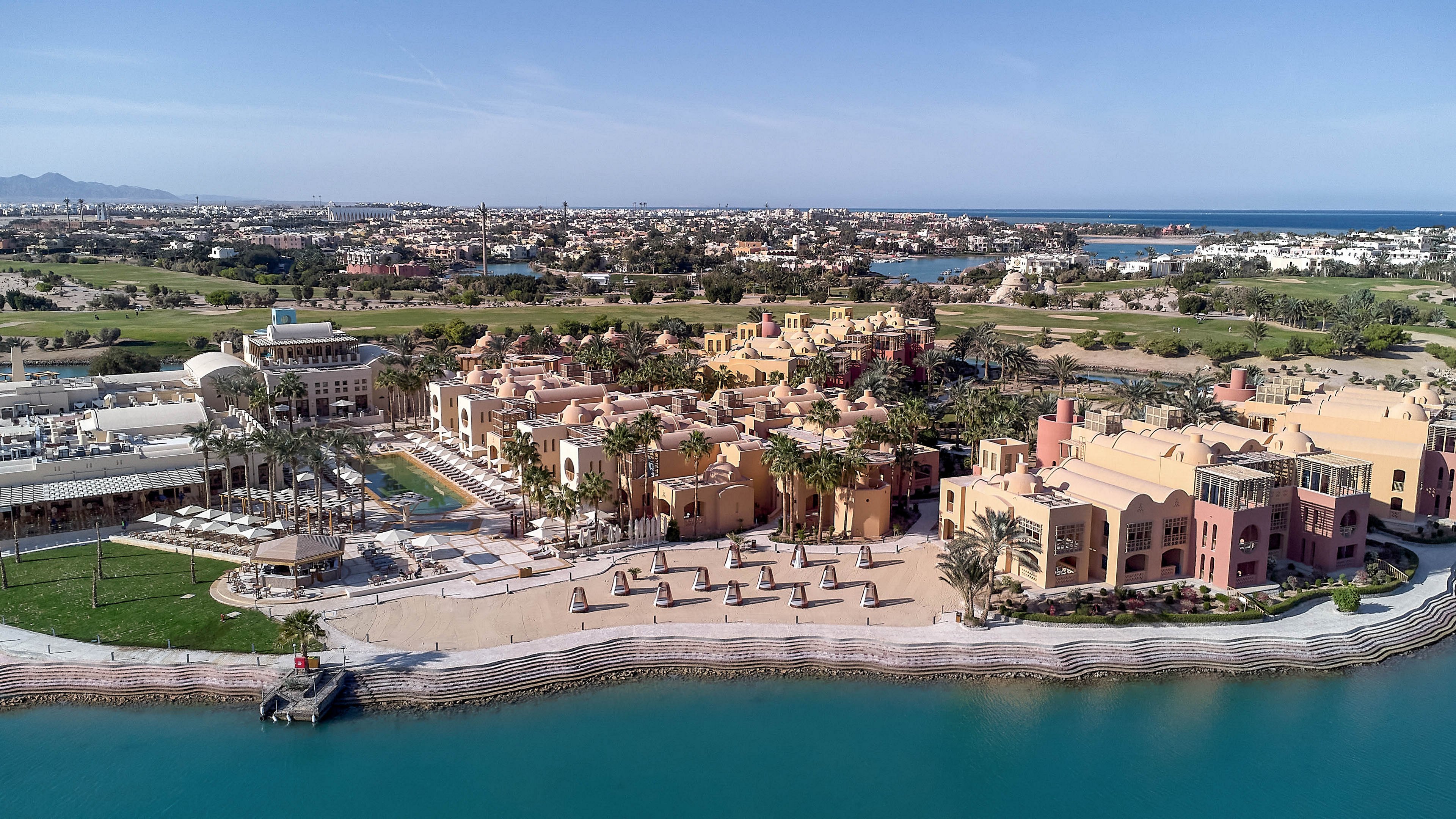 An expansive waterfront resort featuring Mediterranean-style architecture with beige and terracotta tones. The setting includes a calm blue waterway, lush greenery, and a distant view of a golf course and cityscape. The image highlights outdoor seating areas, palm trees, and a serene atmosphere under clear skies.