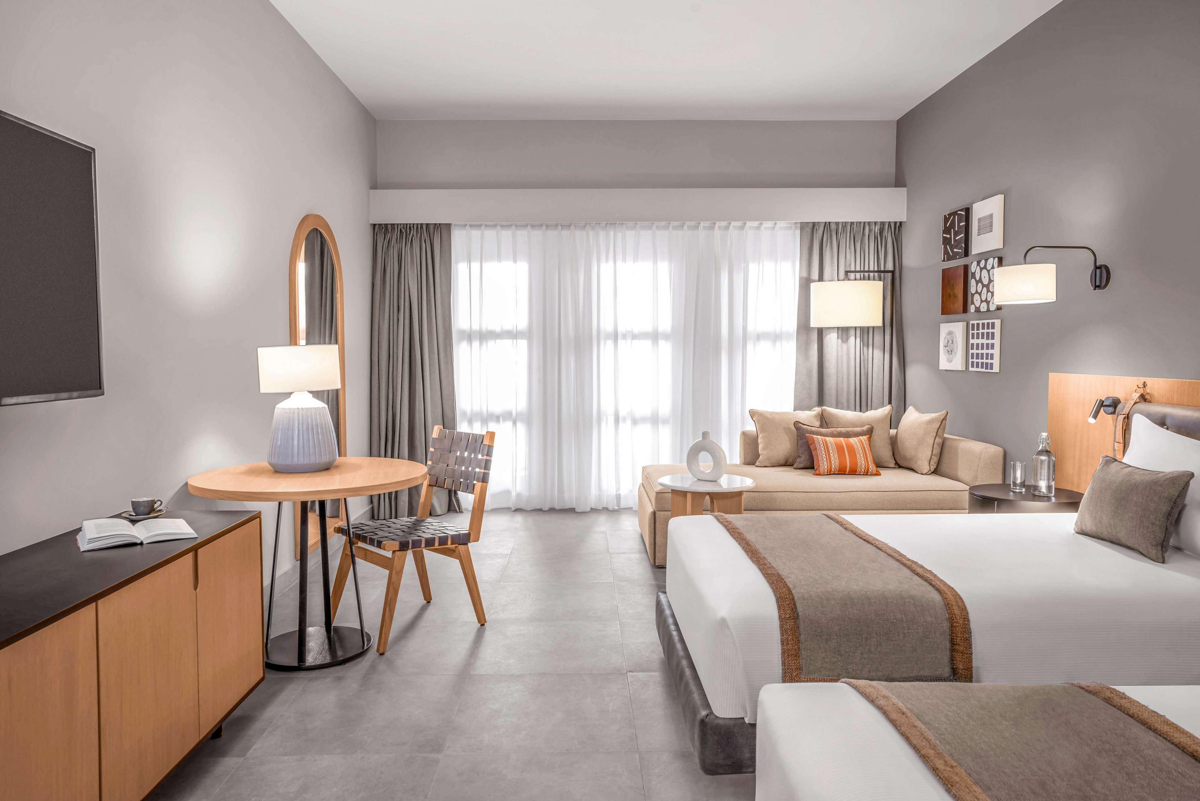 A contemporary hotel room featuring two neatly made twin beds with beige and brown accents. The room includes a seating area with a sofa, a round table with a chair, and a large window with sheer curtains allowing natural light. Neutral tones dominate the decor, creating a calm and inviting atmosphere. A flat-screen TV and decorative elements complete the space.