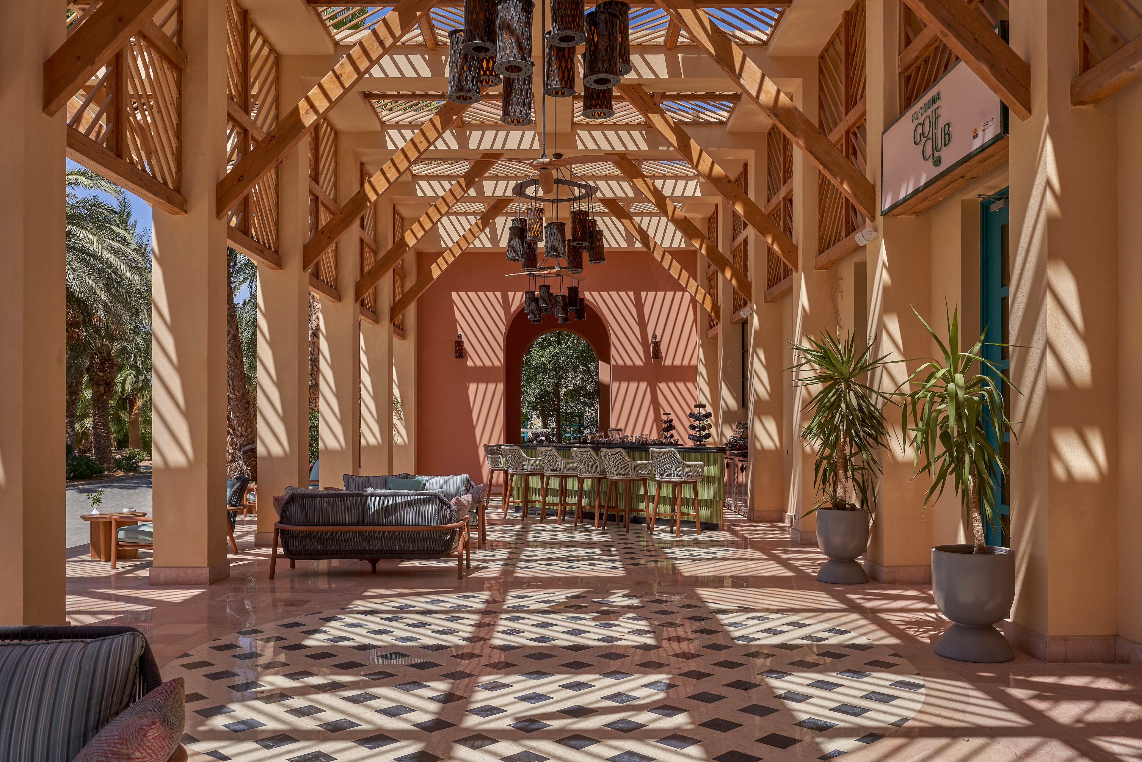 A spacious outdoor seating area featuring wooden beams and patterned tile flooring. The space is adorned with potted plants and stylish furniture, creating a relaxed and inviting atmosphere. Sunlight streams through the slatted roof, casting intricate shadows on the floor. A dining table is visible in the background, surrounded by greenery.