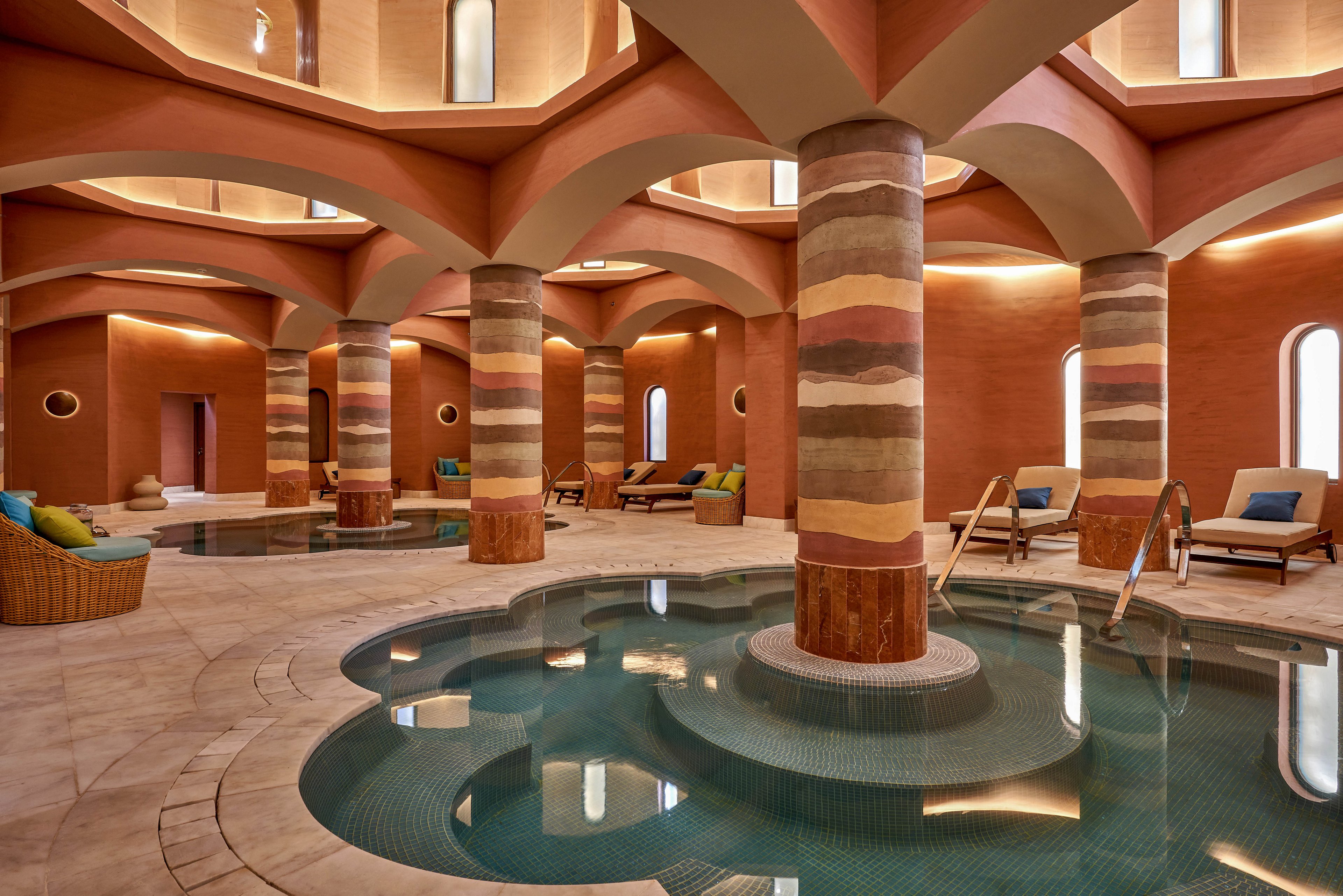 A serene indoor spa featuring multiple pools surrounded by elegant, earthy-toned columns and arches. Comfortable lounge chairs with cushions are arranged around the space, creating a relaxing atmosphere. The warm lighting and natural textures enhance the tranquil ambiance, perfect for unwinding.