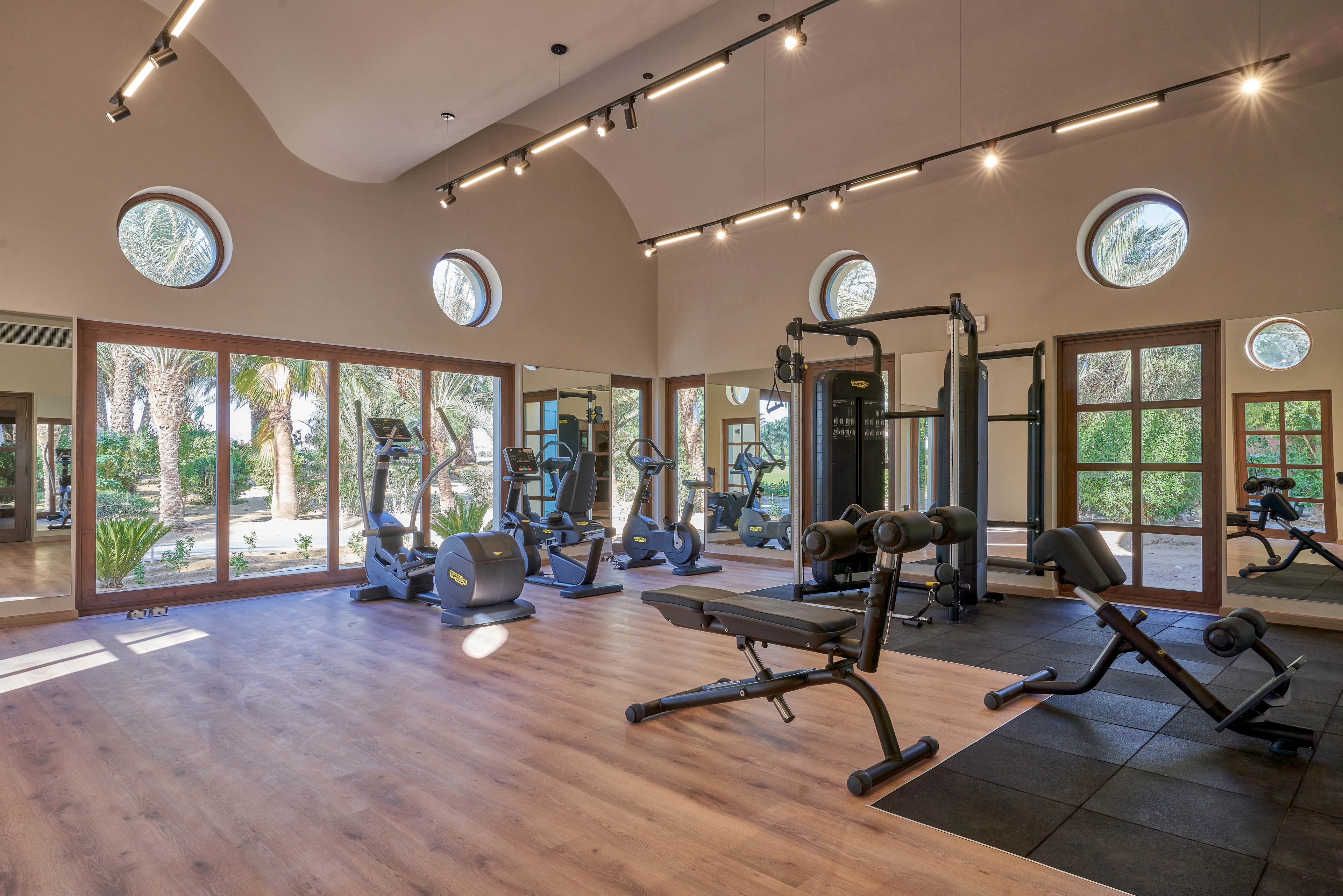 A spacious gym with modern fitness equipment, including treadmills, weight benches, and exercise machines. The room features large windows with views of greenery and circular skylights for natural lighting. The wooden flooring and neutral tones create a welcoming and professional atmosphere.