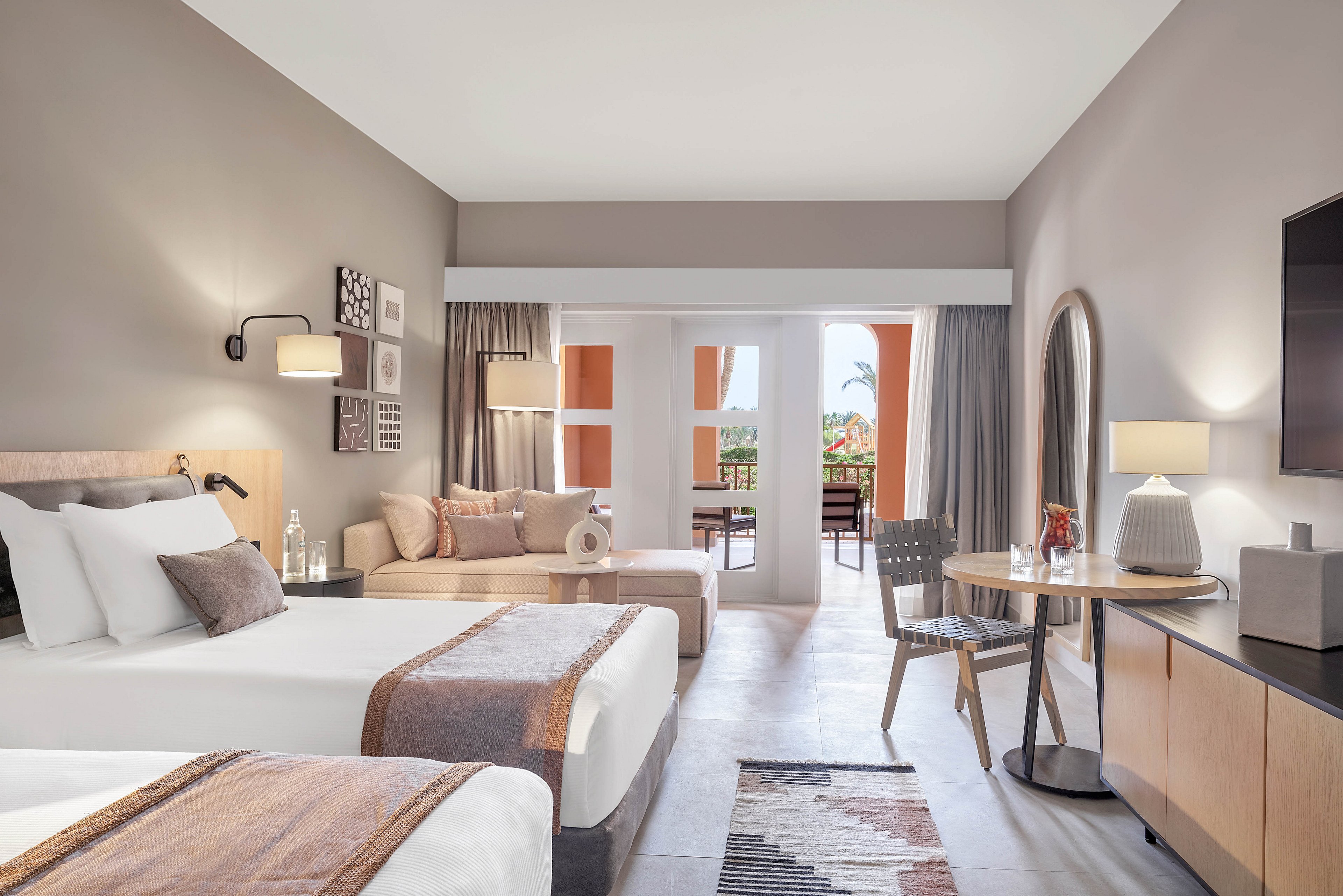 A contemporary hotel room featuring two neatly made twin beds with neutral-toned bedding. The room includes a seating area, a round table with chairs, and a balcony overlooking an outdoor view. Warm lighting and minimalist decor create a cozy and inviting atmosphere. A patterned rug and decorative accents add a touch of style.