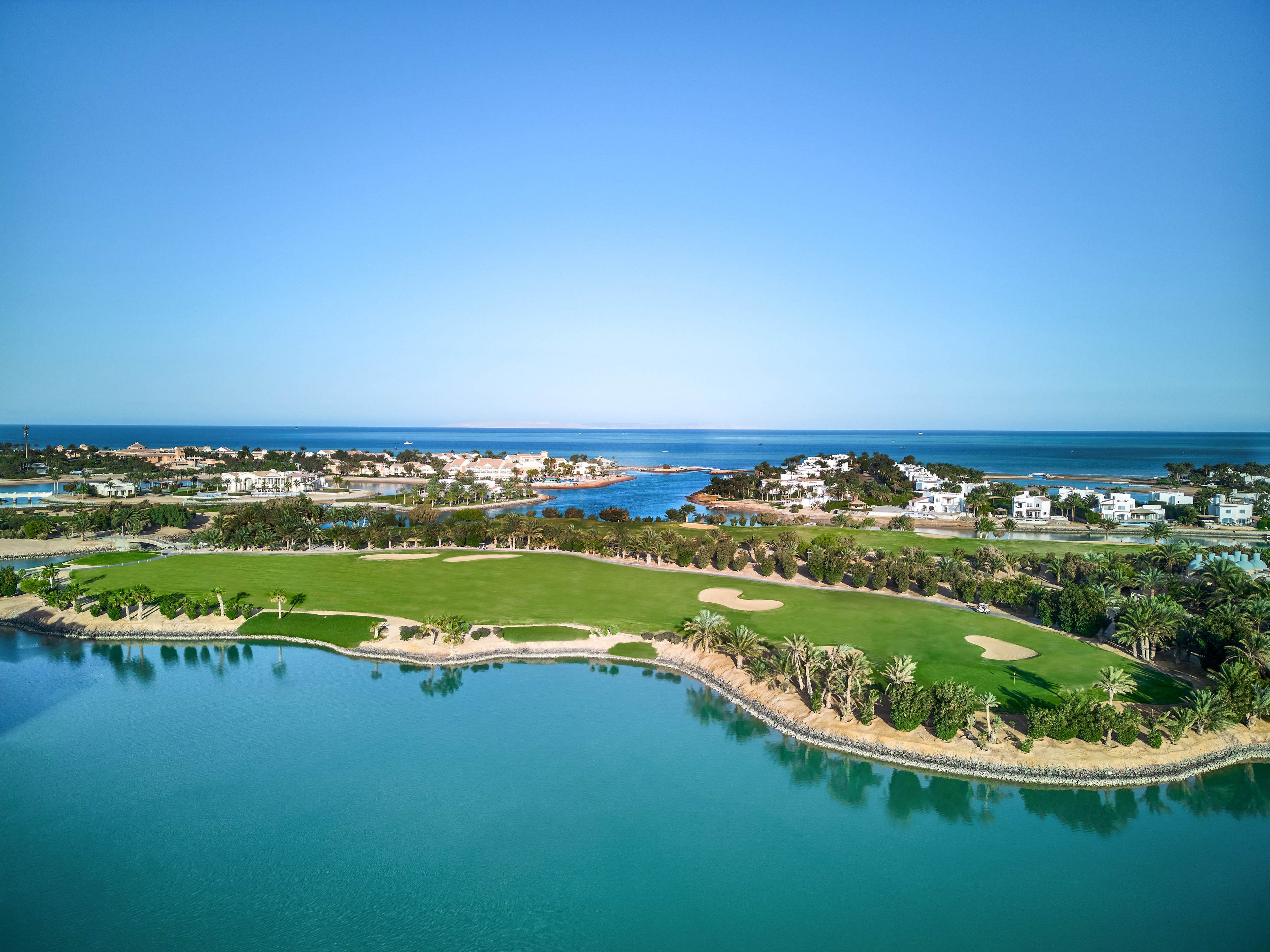 Aerial view of a lush green golf course surrounded by a serene lagoon and coastal landscape. The setting features clear blue skies, calm waters, and luxurious residential properties in the background. The vibrant greenery contrasts beautifully with the tranquil blue tones of the water and sky.