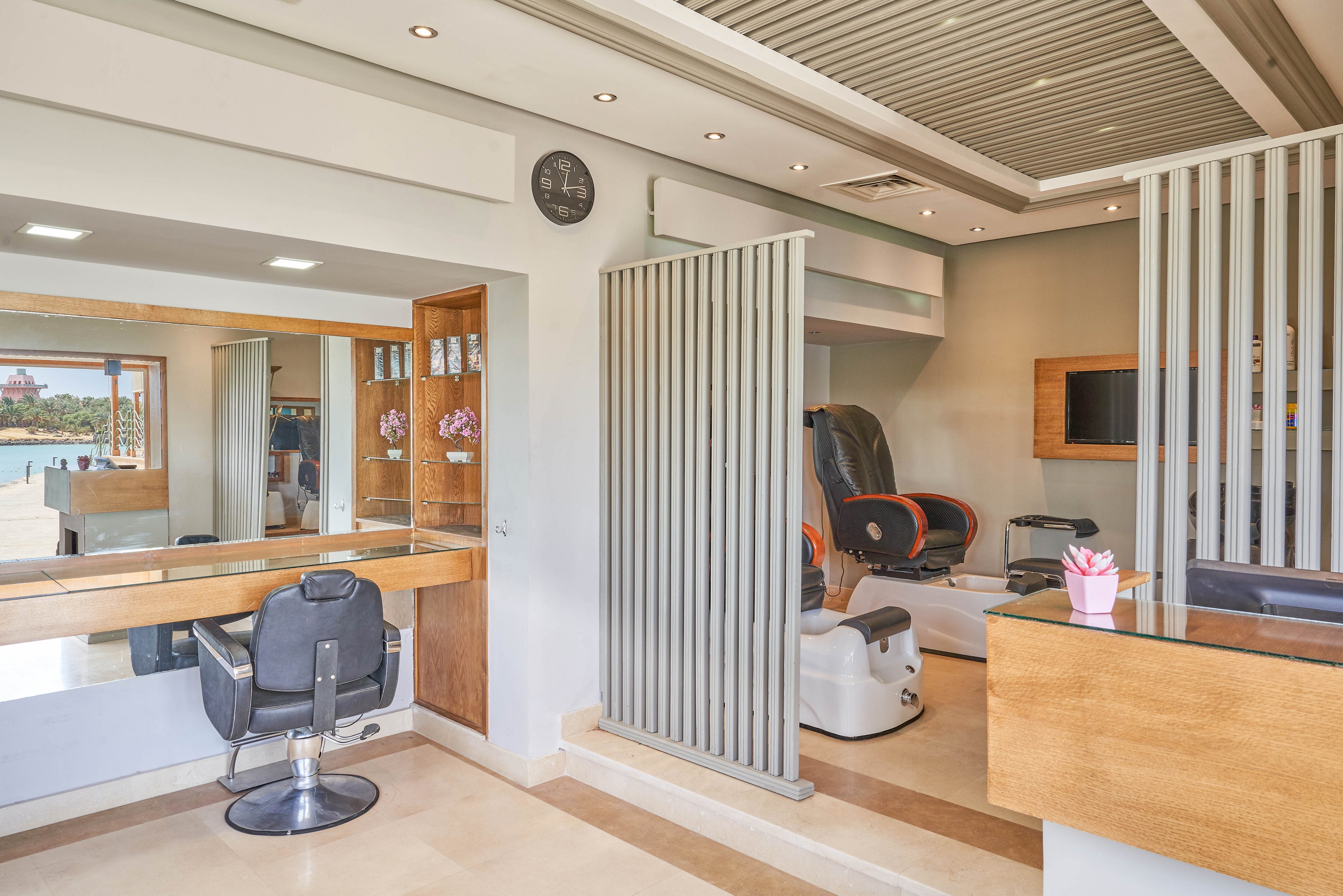 A contemporary salon featuring a sleek design with wooden and metallic accents. The space includes a styling station, a massage chair, and a reception desk. Large windows provide a scenic view of a lake and outdoor area. A wall clock is visible, adding functionality to the stylish setting.