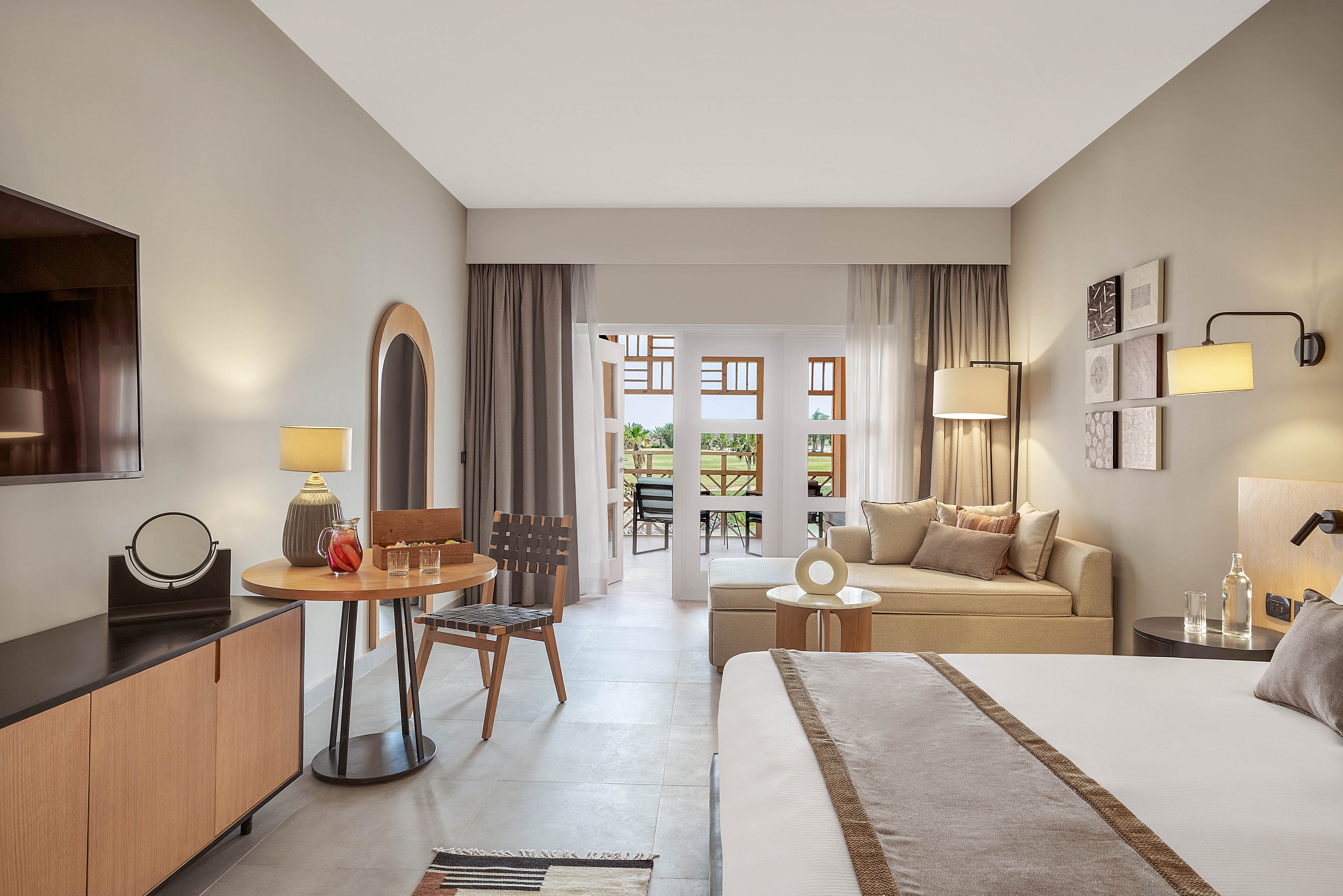 A spacious and elegantly designed hotel room featuring a comfortable bed, a cozy seating area, and a work desk. The room is decorated in neutral tones with modern furnishings and opens to a balcony with outdoor seating and a scenic view. Warm lighting and minimalist decor create a relaxing ambiance.