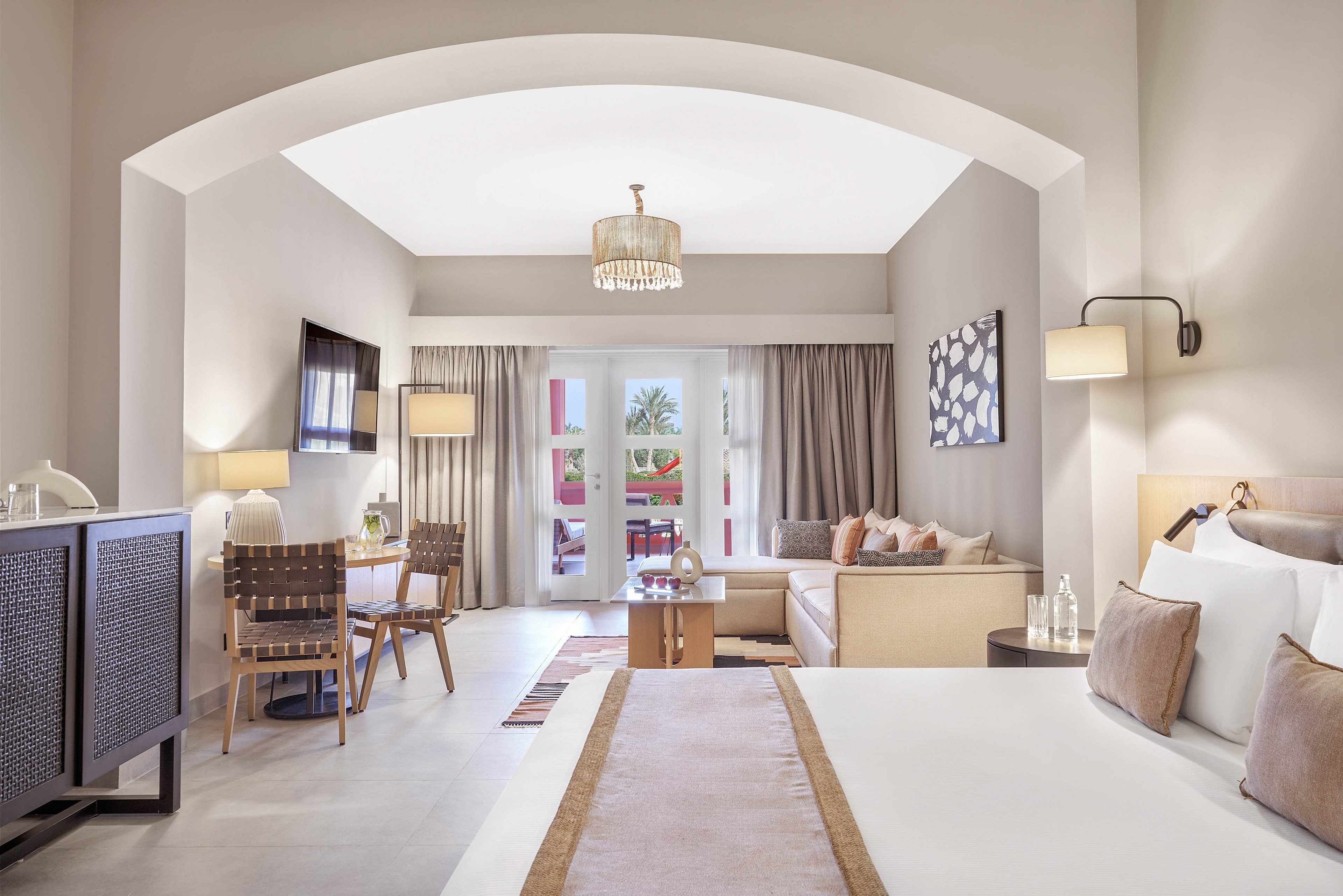 A spacious and elegantly designed hotel room featuring a large bed, seating area, and a balcony with outdoor views. Neutral tones and soft lighting create a warm and inviting atmosphere. The room includes a flat-screen TV, wooden furniture, and decorative accents.