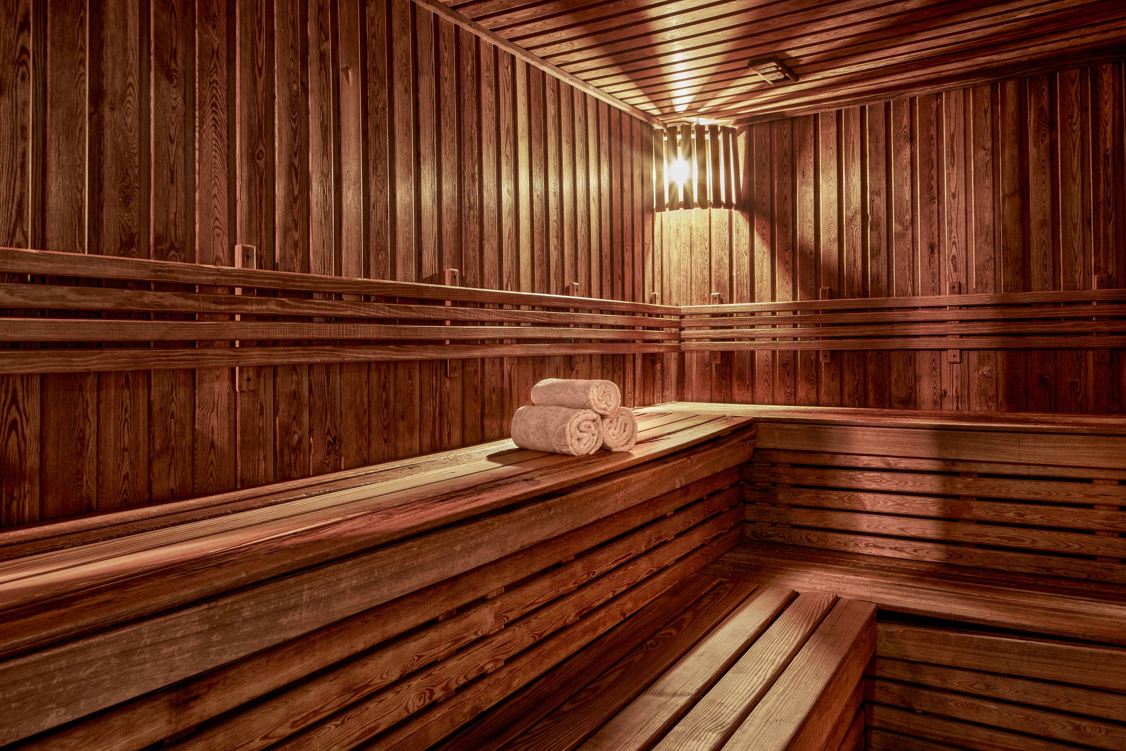 A warm and inviting wooden sauna interior featuring neatly rolled white towels on a bench. The space is illuminated by soft, ambient lighting from a wall-mounted lamp. The wooden panels and benches create a cozy and relaxing atmosphere, ideal for unwinding and rejuvenation.
