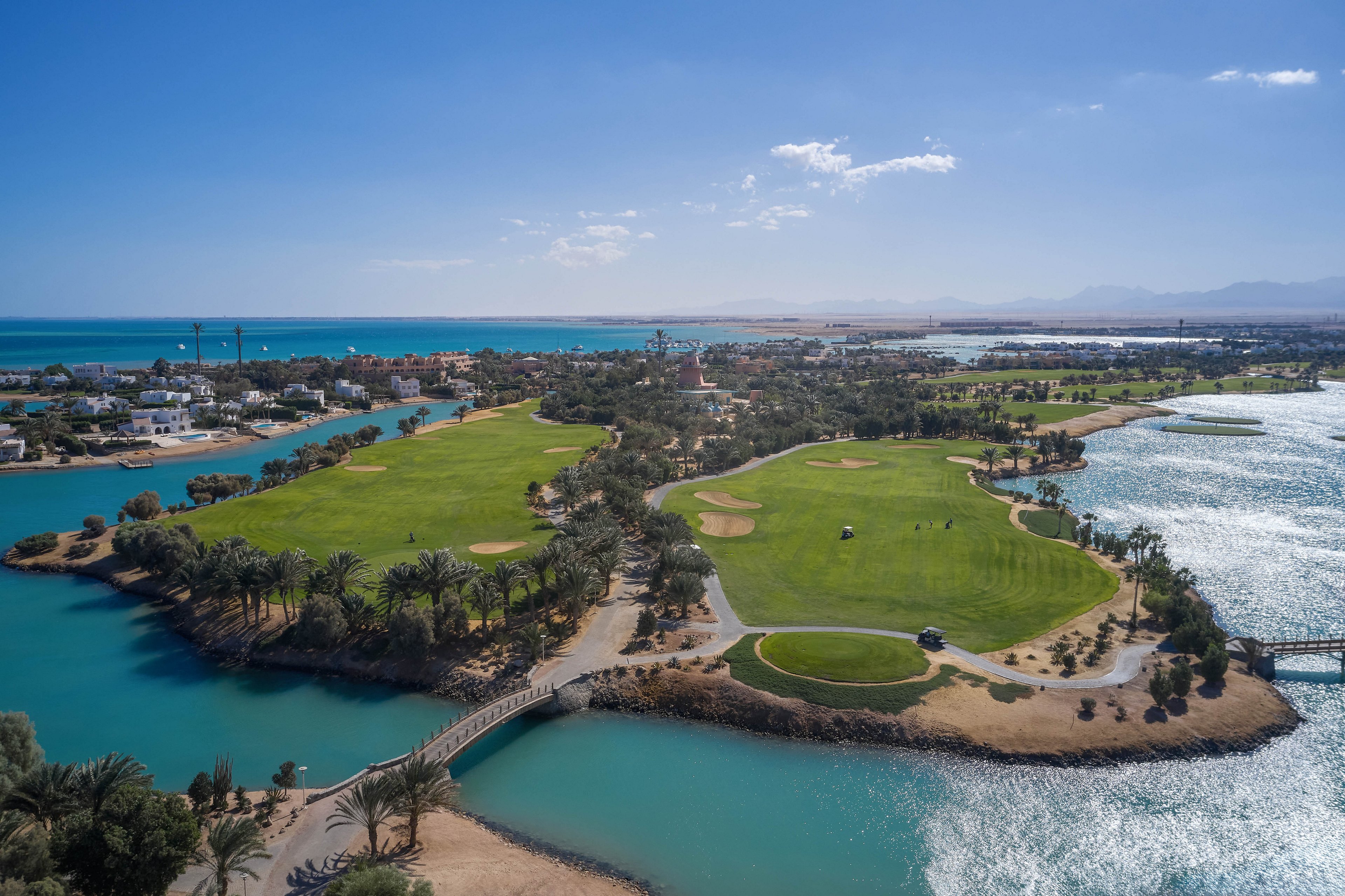 An expansive golf course surrounded by lush greenery and water bodies, located near a coastal area. The image showcases a sunny day with clear skies and a serene landscape. A small bridge connects parts of the course, adding to the scenic beauty.