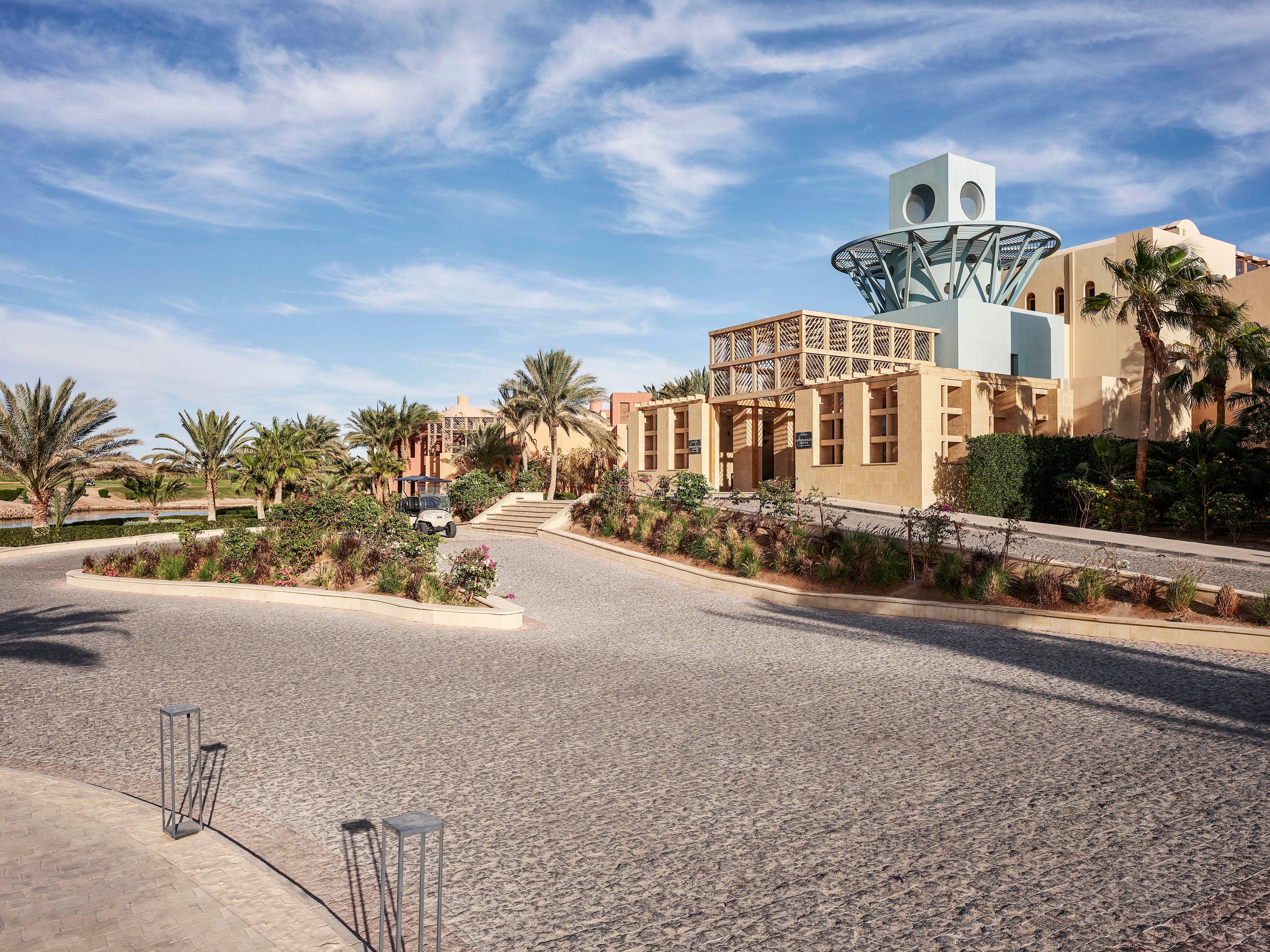 A contemporary building with unique architectural design, featuring a circular structure on top. The setting includes a paved road, landscaped greenery, and palm trees under a clear blue sky. The scene exudes a calm and sunny atmosphere.