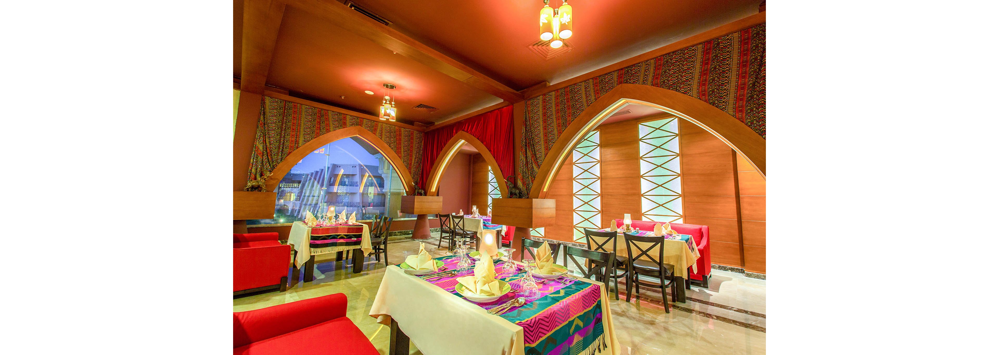 A vibrant restaurant dining area features arched windows and decorative textiles on the walls. Tables are set with folded napkins, glassware, and colorful tablecloths, creating a welcoming atmosphere. The lighting is warm, and the seating includes both red sofas and black chairs. No visible text or numbers are present in the image.