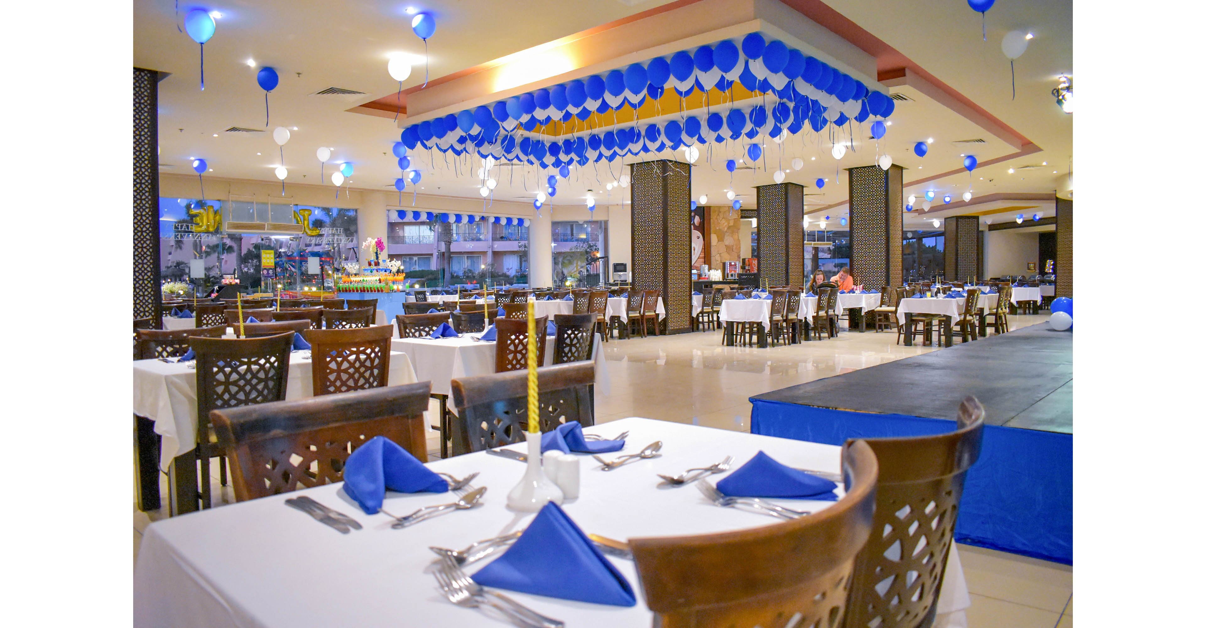 A spacious restaurant dining area is set up with neatly arranged tables covered in white cloths and blue napkins. The ceiling is adorned with numerous blue and white balloons, creating a festive atmosphere. The setting appears ready for a celebration or special event, with cutlery and glasses meticulously placed. The overall mood is cheerful and inviting, with bright lighting and a clean, modern interior.