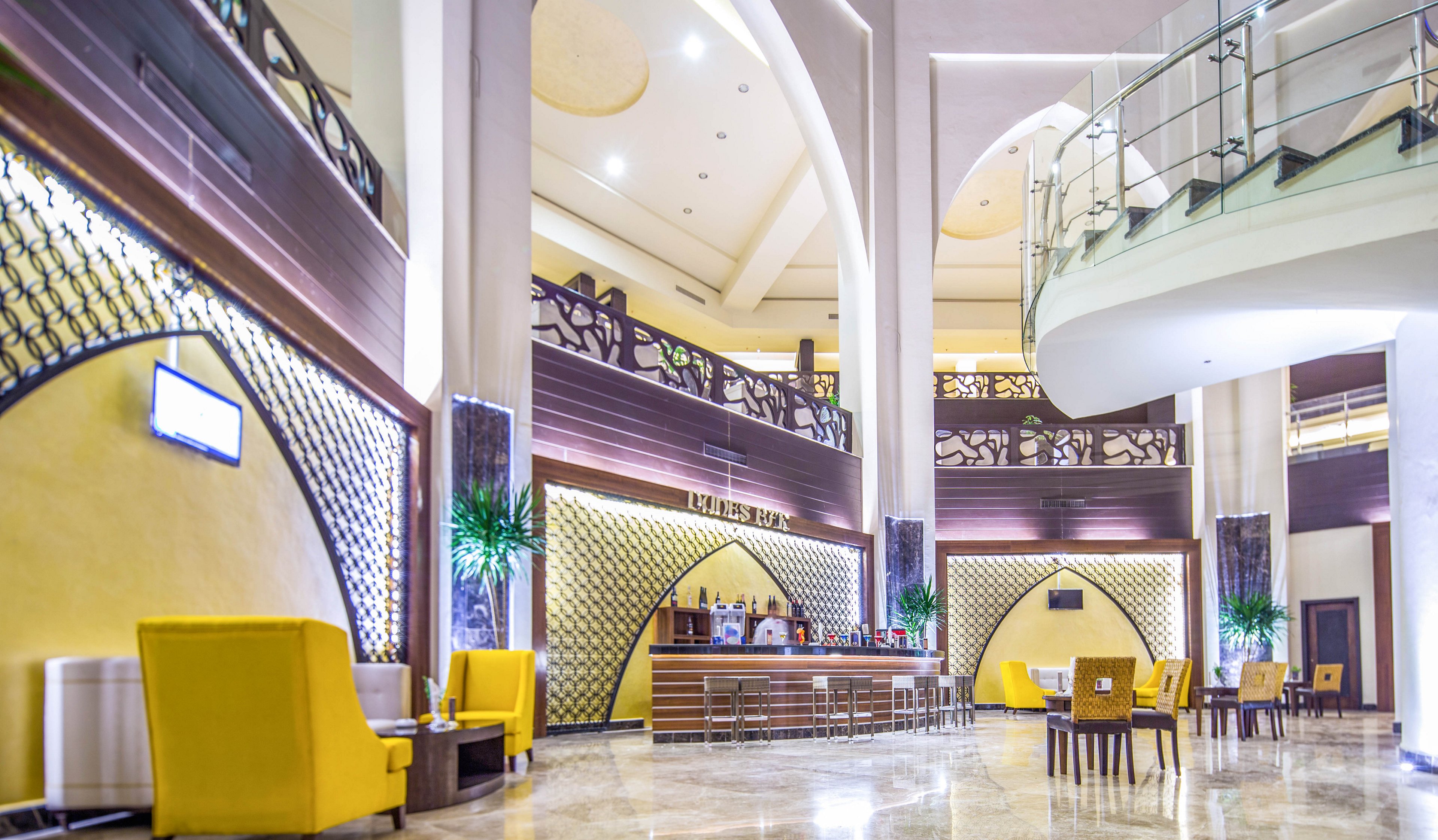 A spacious hotel lobby features a central bar area with high stools and decorative arch designs. Bright yellow armchairs and sofas are arranged in seating areas, complemented by tall indoor plants and contemporary lighting. The setting is airy and elegant, with a neutral palette accented by yellow and dark wood tones. A staircase with glass railing leads to an upper level, and digital screens are visible on the walls.