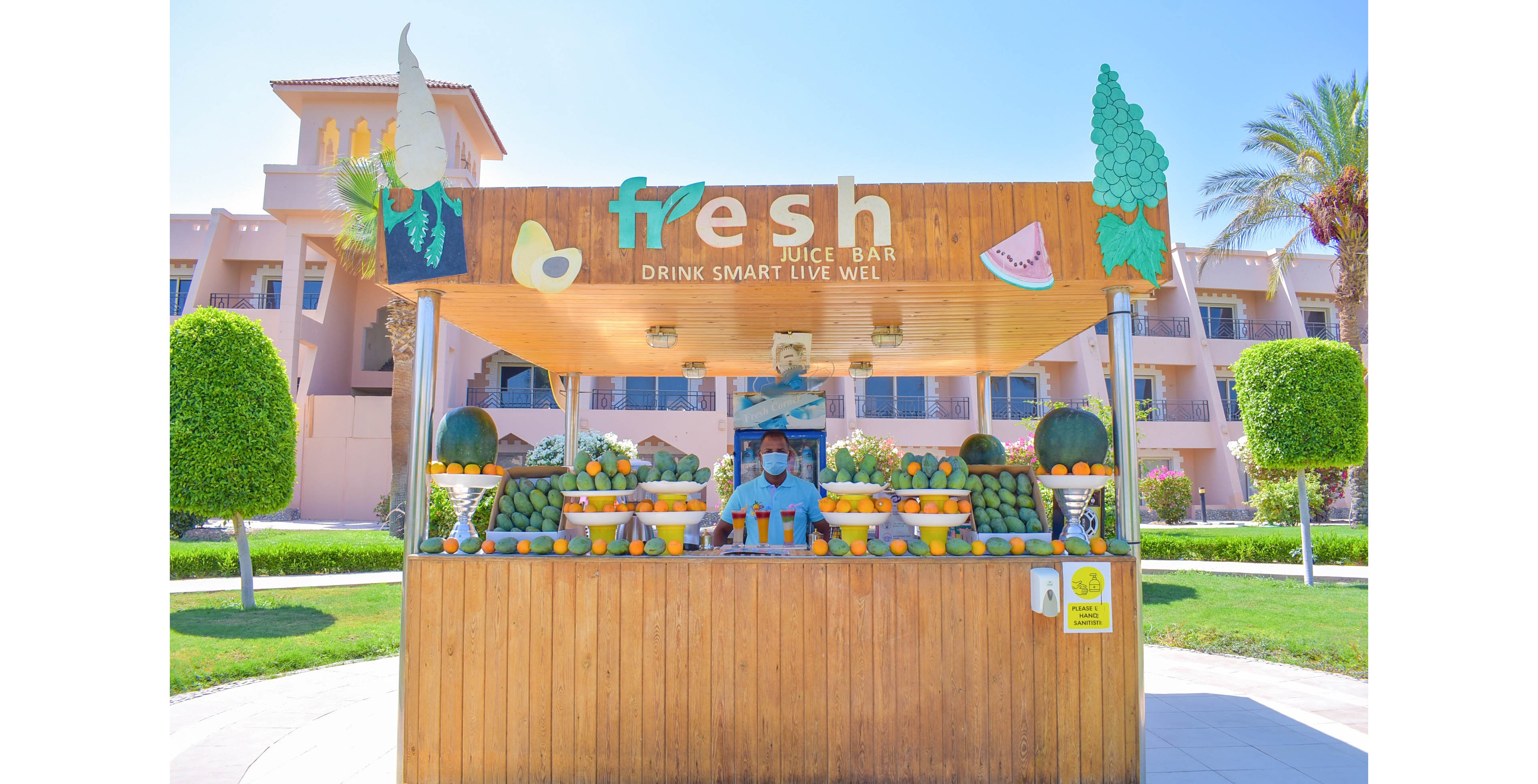 A wooden juice bar stand labeled 'fresh juice bar' is set outdoors in a sunny, landscaped area. The stand is decorated with various fruits including oranges, lemons, and watermelons, and a masked vendor stands behind the counter. The sign above reads 'DRINK SMART. LIVE WELL' and features fruit illustrations, with a clear blue sky and palm trees in the background.