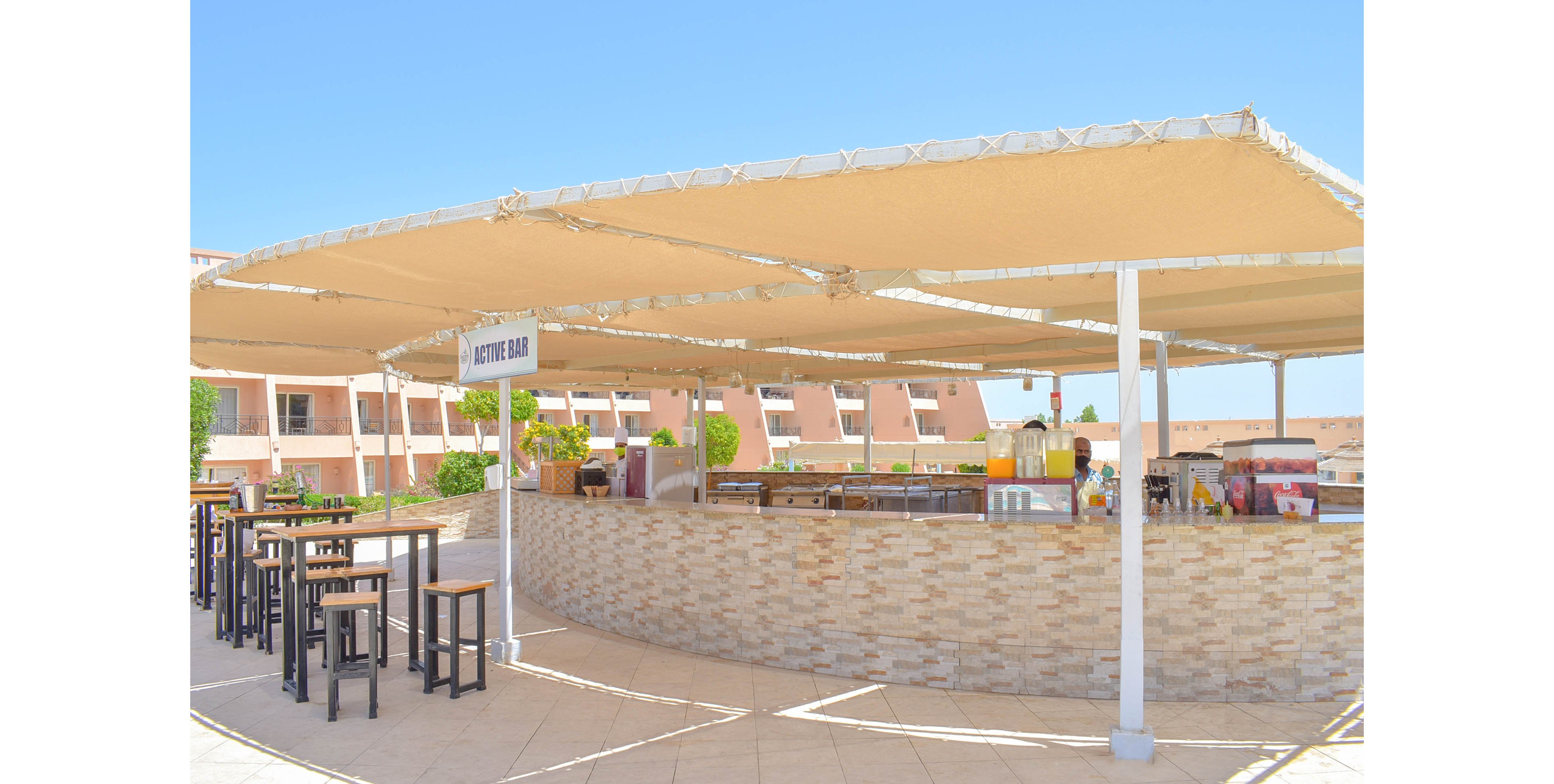 An open-air juice bar is situated on a tiled patio, featuring a curved counter with various beverage dispensers and supplies. The area is shaded by large canvas canopies, with high tables and stools arranged nearby. The setting is bright and sunny, with a clear blue sky and hotel buildings in the background. A sign reading 'JUICE BAR' is clearly visible above the counter.