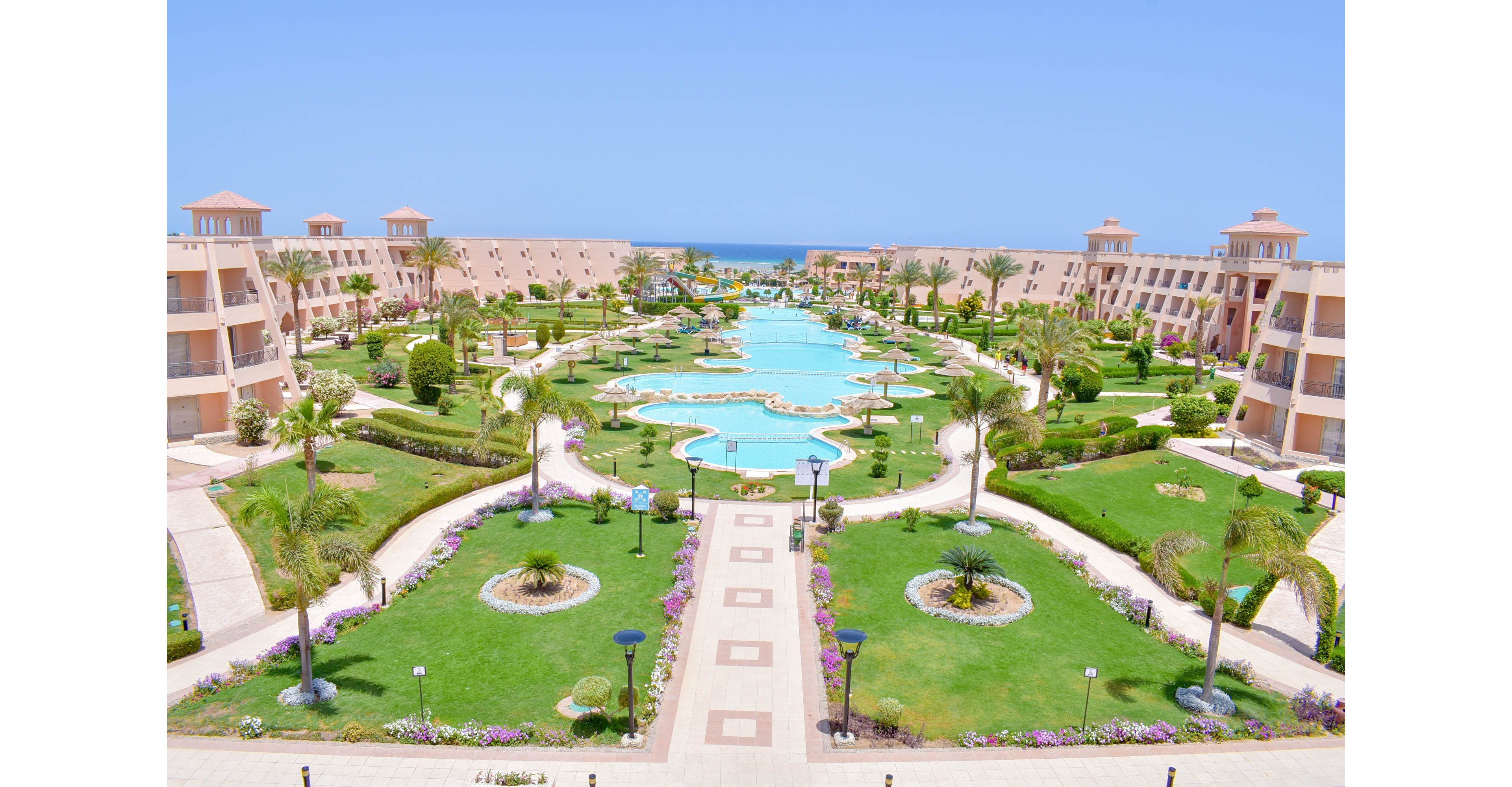 A spacious resort complex features symmetrical buildings flanking a large, winding swimming pool at its center. The scene is set outdoors under a clear blue sky, with manicured lawns, palm trees, and landscaped gardens enhancing the tranquil atmosphere. Pathways and decorative lighting line the grounds, leading toward the distant sea visible on the horizon. The architecture is modern, with light-colored facades and balconies overlooking the pool area. No visible people or text are present in the image.