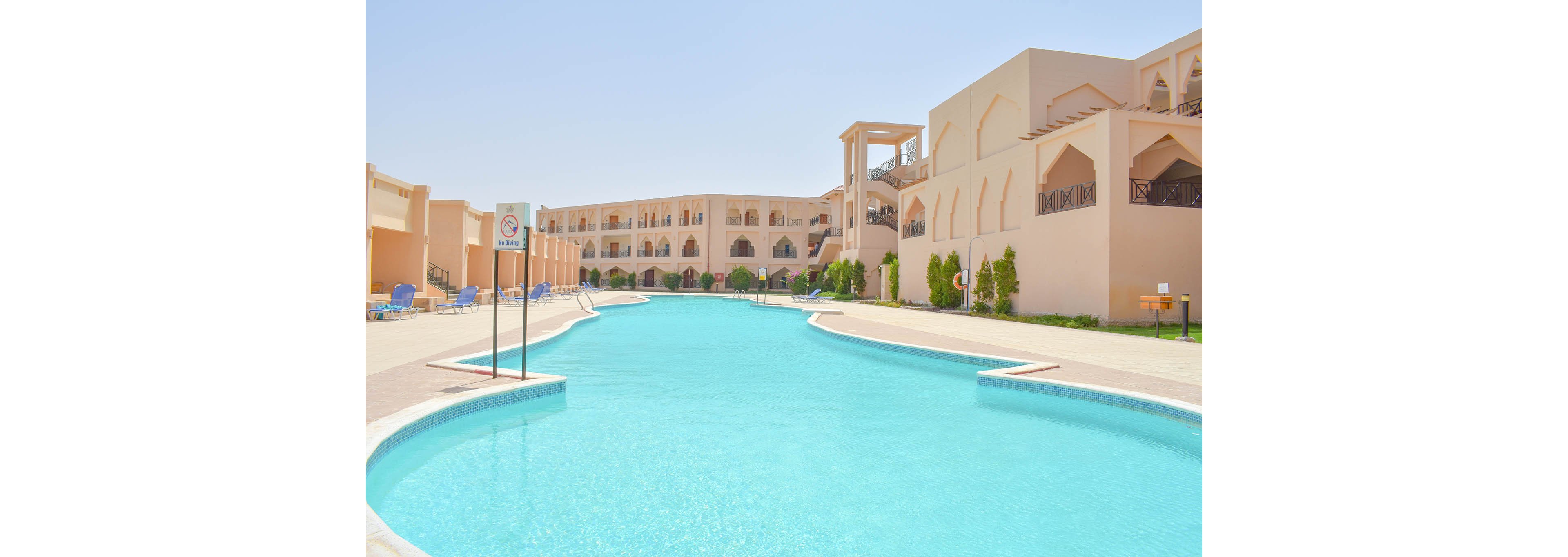 A large, clear swimming pool stretches between beige Mediterranean-style resort buildings under a bright blue sky. The setting is tranquil and sunlit, with lounge chairs and greenery lining the poolside. A sign is visible near the pool, and the overall mood is relaxed and inviting, ideal for vacation or leisure activities.
