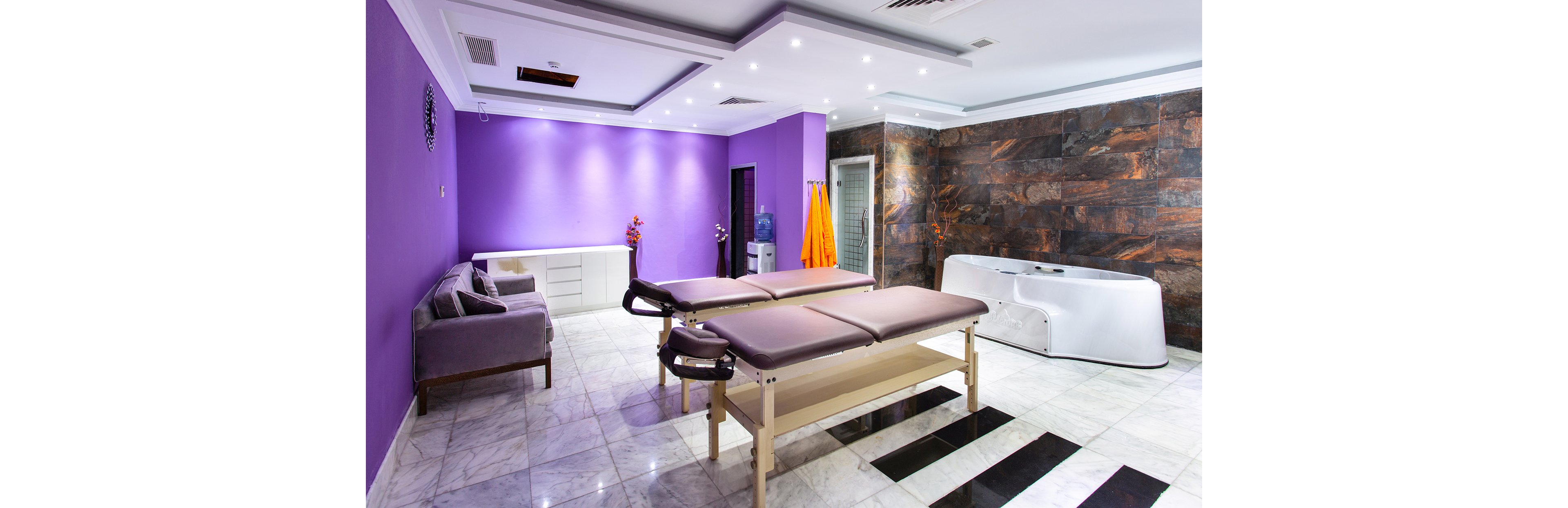 A contemporary spa room features two massage tables at the center, set against a vibrant purple accent wall. The space includes a sleek sofa, a white storage unit, and a modern hydrotherapy tub. Bright lighting and marble flooring create a clean, inviting atmosphere, with orange towels adding a pop of color. No visible text or numeric values are present in the image.