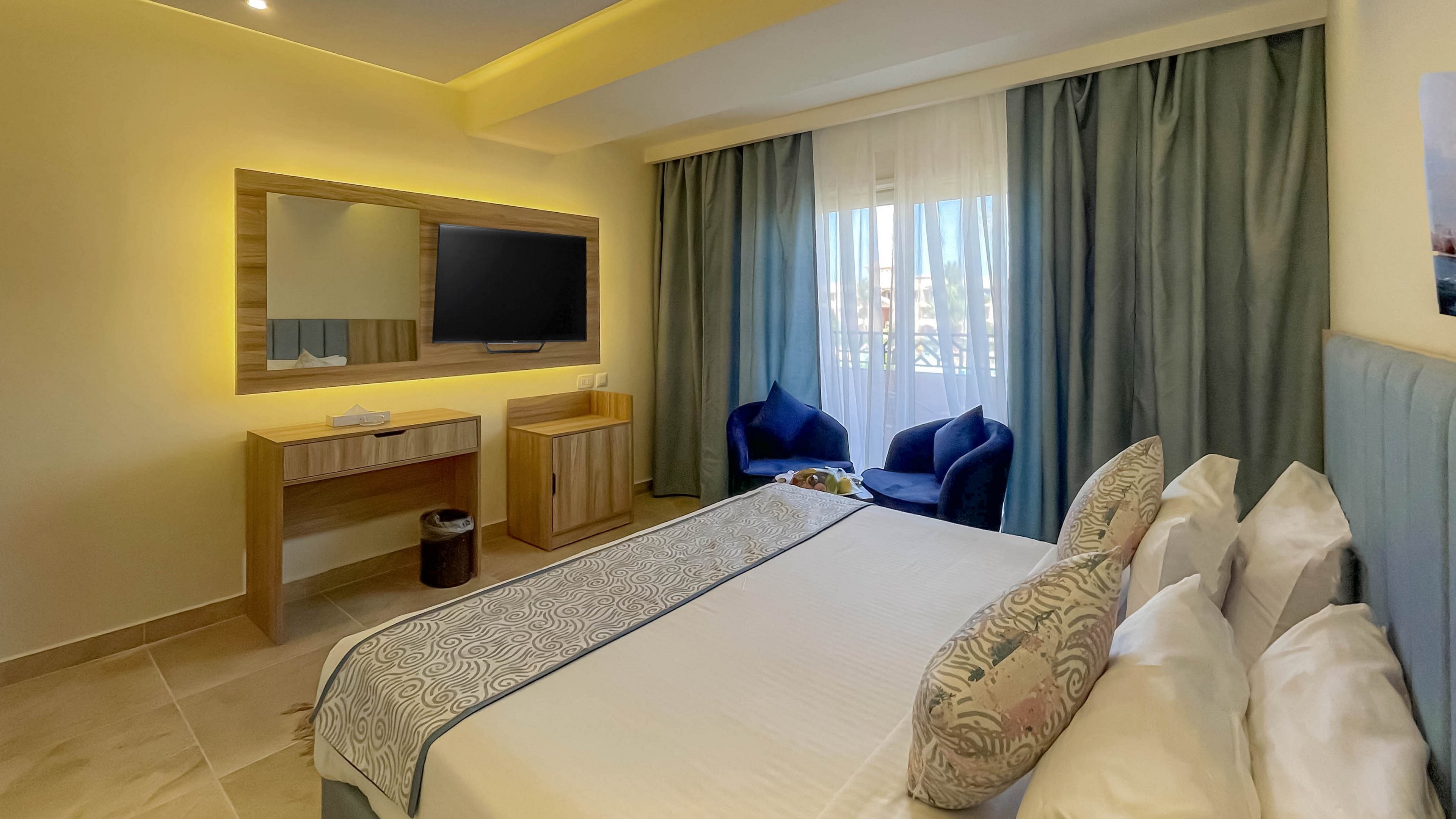 A contemporary hotel room features a neatly made bed with decorative pillows, a wall-mounted flat-screen television, and a wooden vanity with a mirror. The room is well-lit with natural light streaming through large windows covered by sheer curtains and blue drapes. Two dark blue armchairs and a small table are positioned near the window, creating a cozy seating area.
