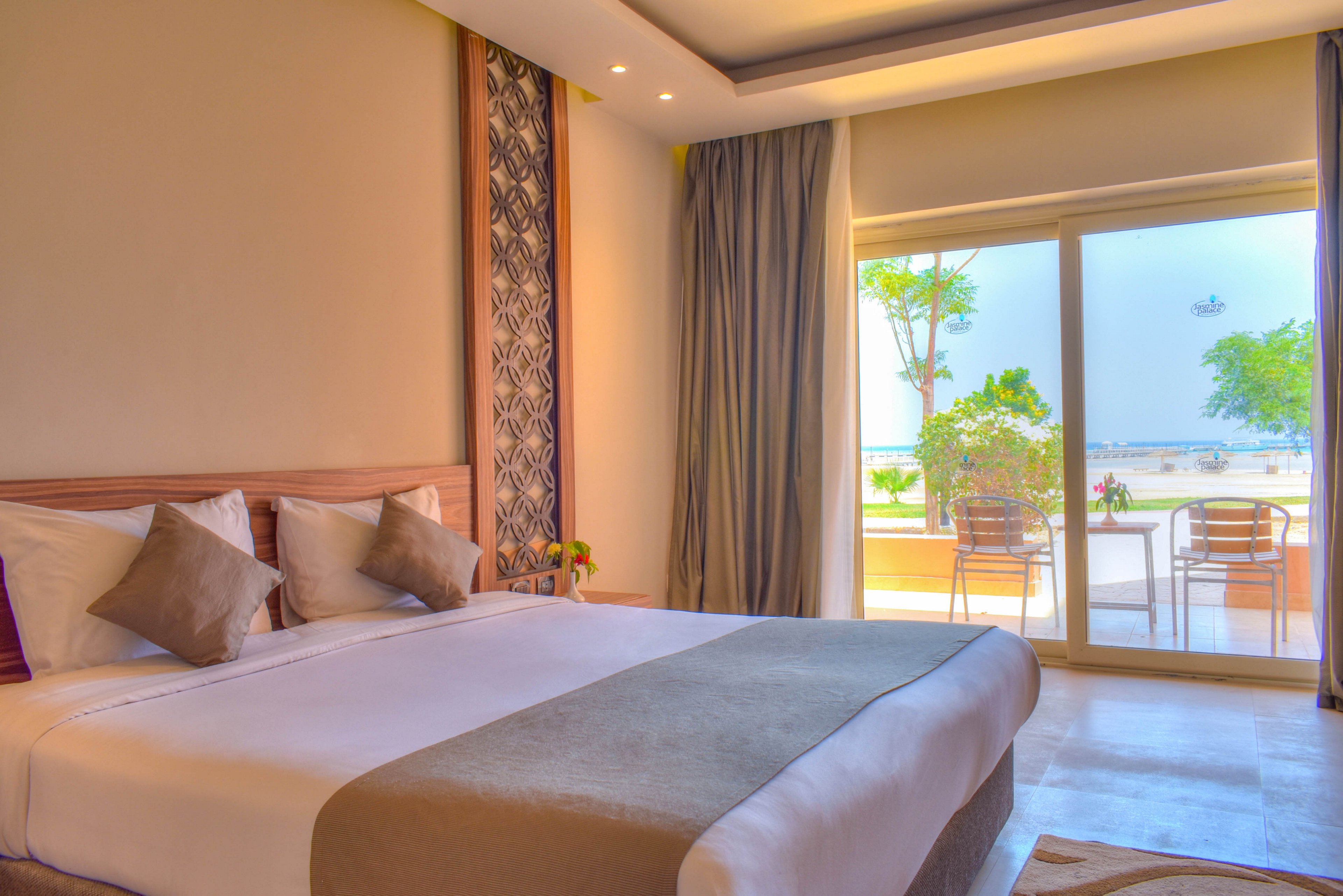 A spacious hotel room features a large bed with neutral bedding and decorative pillows. The room opens to a private patio with outdoor seating, overlooking a serene beach and ocean. Soft natural light fills the space, highlighting the contemporary decor and tranquil atmosphere.