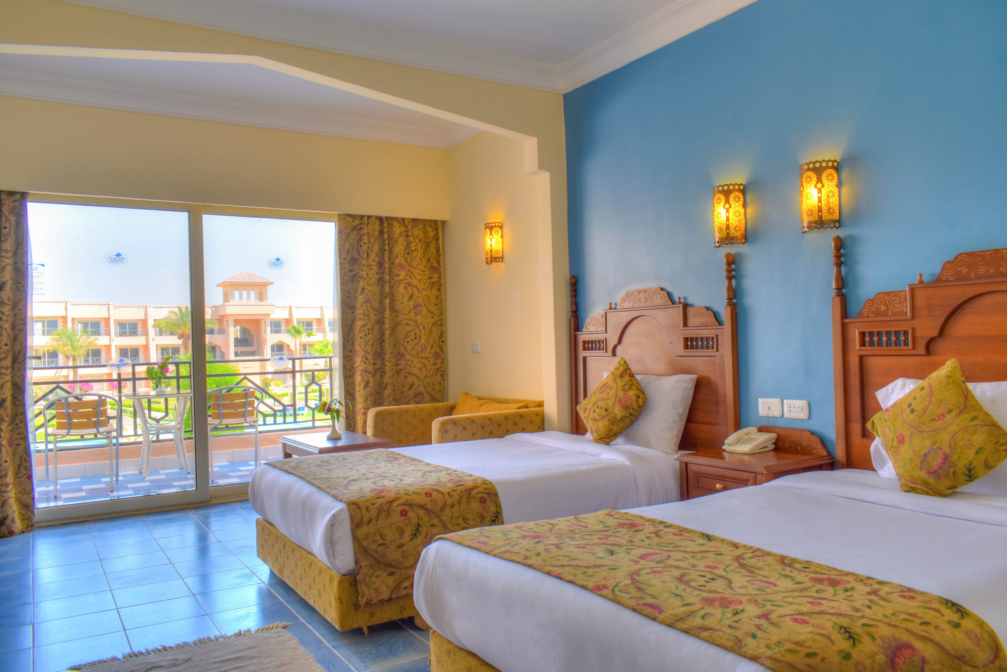 A spacious hotel room features two twin beds with ornate wooden headboards and patterned bedding. The room is decorated in warm tones with a blue accent wall and golden lighting fixtures. Large glass doors open to a balcony overlooking a resort courtyard, creating an inviting and airy atmosphere. No visible text or numbers are present in the image.