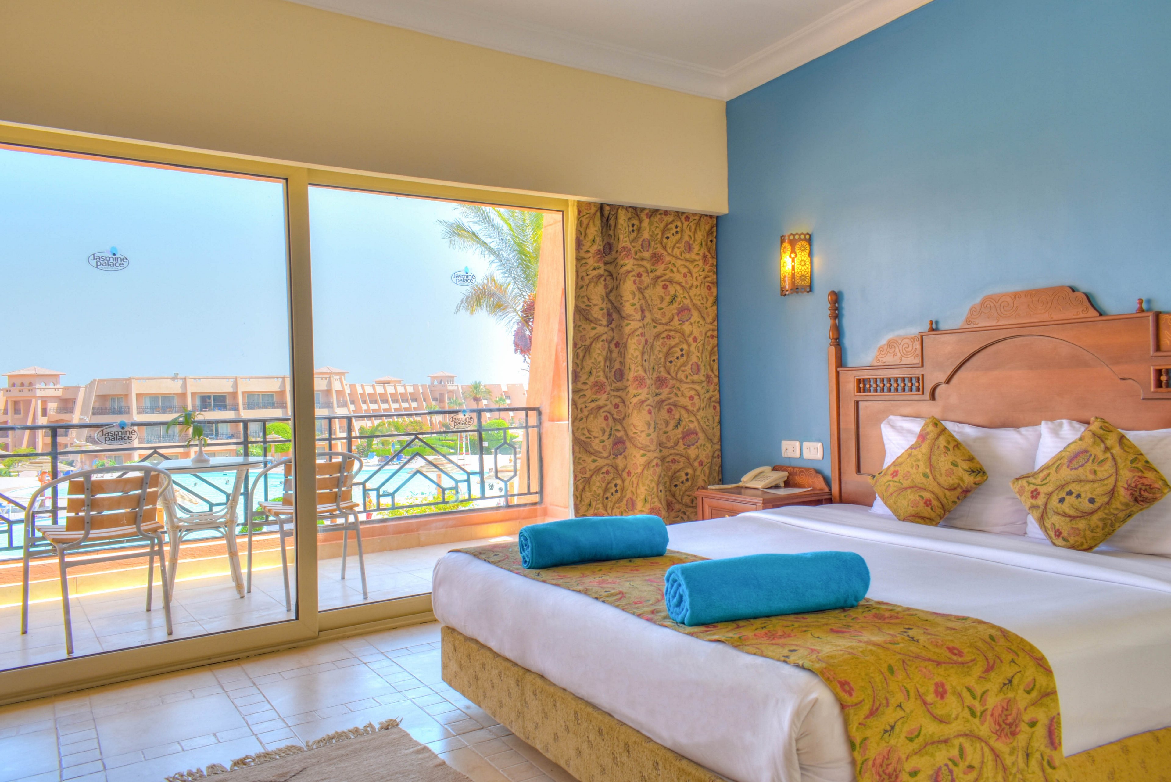 A spacious hotel room features a large bed with patterned pillows and two rolled blue towels. The room opens onto a balcony with chairs, overlooking a pool and palm trees. Warm yellow and blue tones create a welcoming, relaxed atmosphere, with natural light streaming through the glass doors.