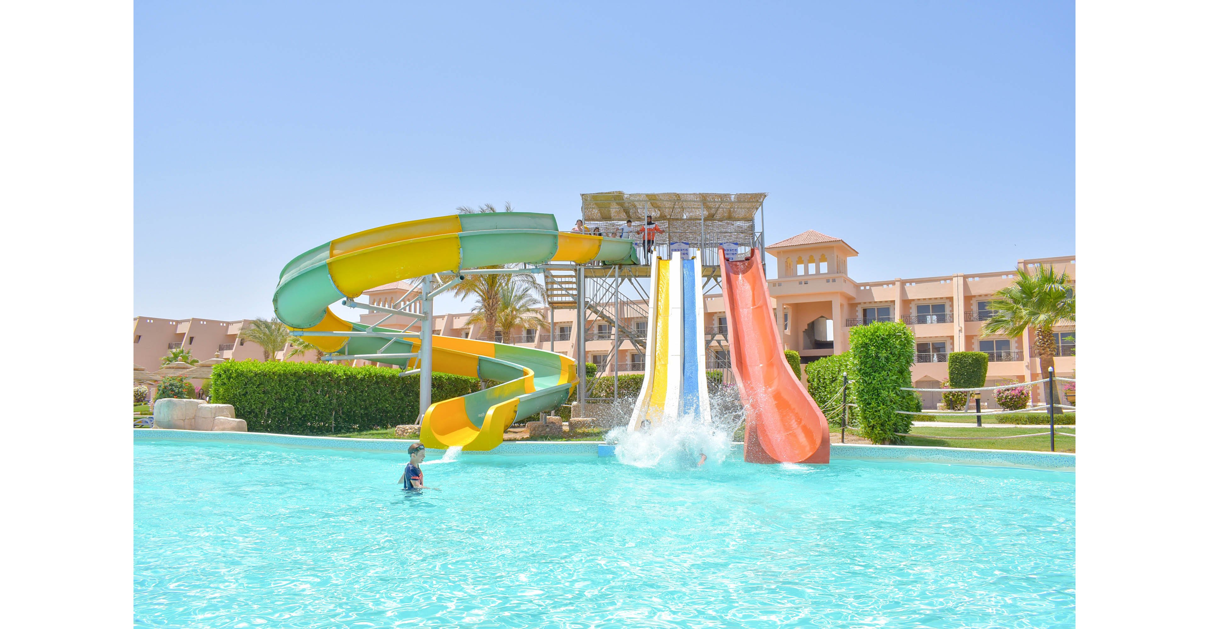 A vibrant water park featuring colorful slides leading into a large outdoor pool. The setting is sunny with clear blue skies, surrounded by lush greenery and a resort-style building in the background. A child is seen enjoying the pool, adding a lively touch to the scene.