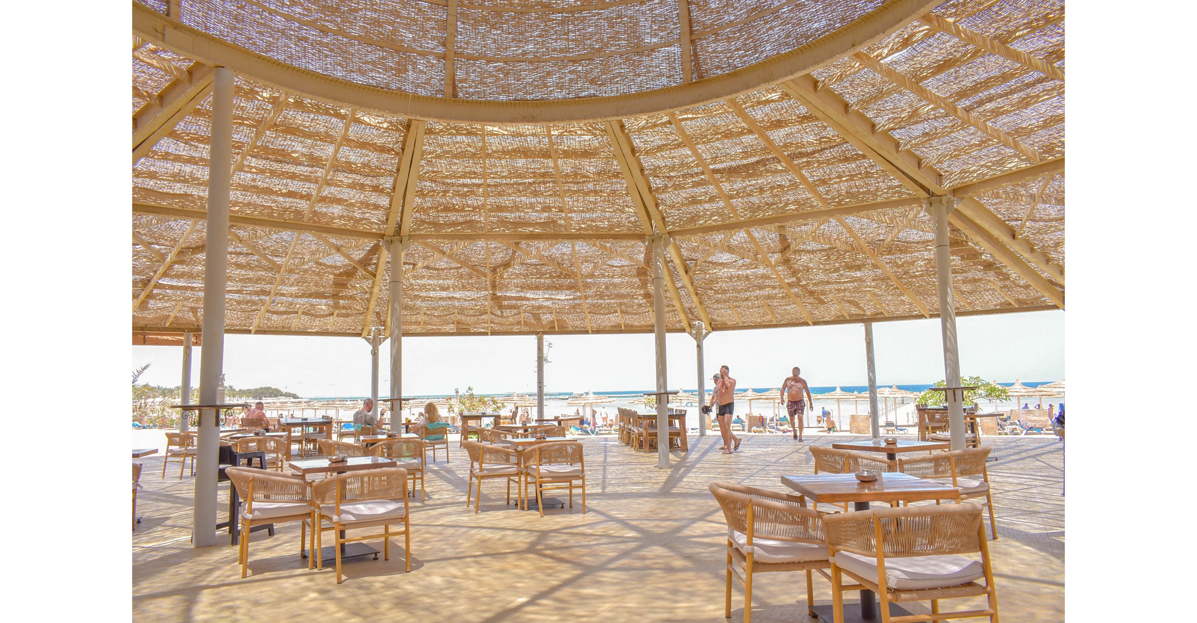 A spacious open-air cafe with a straw-thatched roof, offering a shaded retreat near the beach. Wooden tables and chairs are arranged under the structure, with people enjoying the relaxed atmosphere. The ocean and sandy beach are visible in the background, enhancing the tropical vibe.