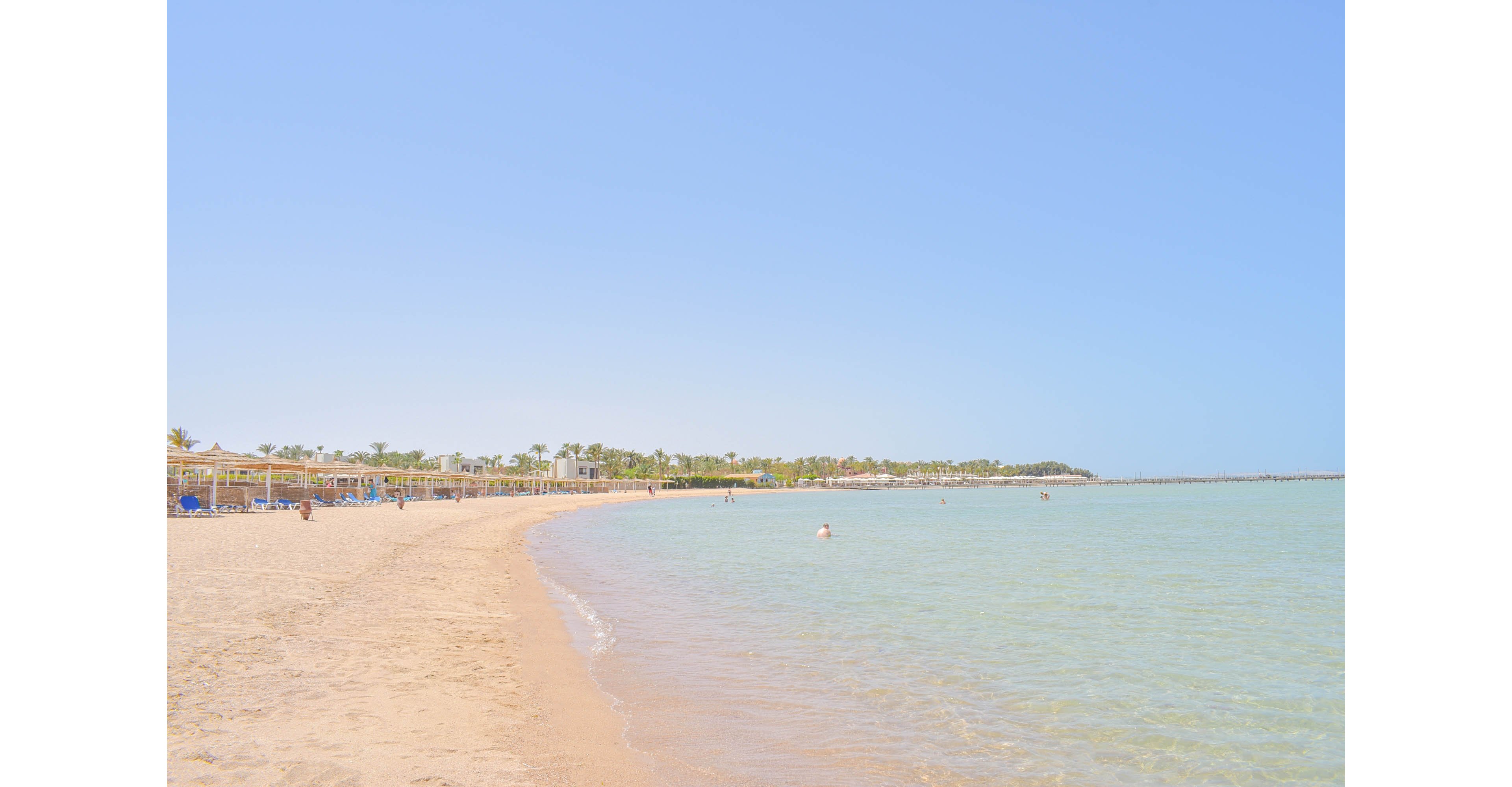 A serene beach scene featuring clear turquoise waters and a sandy shoreline under a bright blue sky. Sun umbrellas and lounge chairs are visible along the shore, with a few people enjoying the water. The distant greenery adds a tropical vibe to the setting.