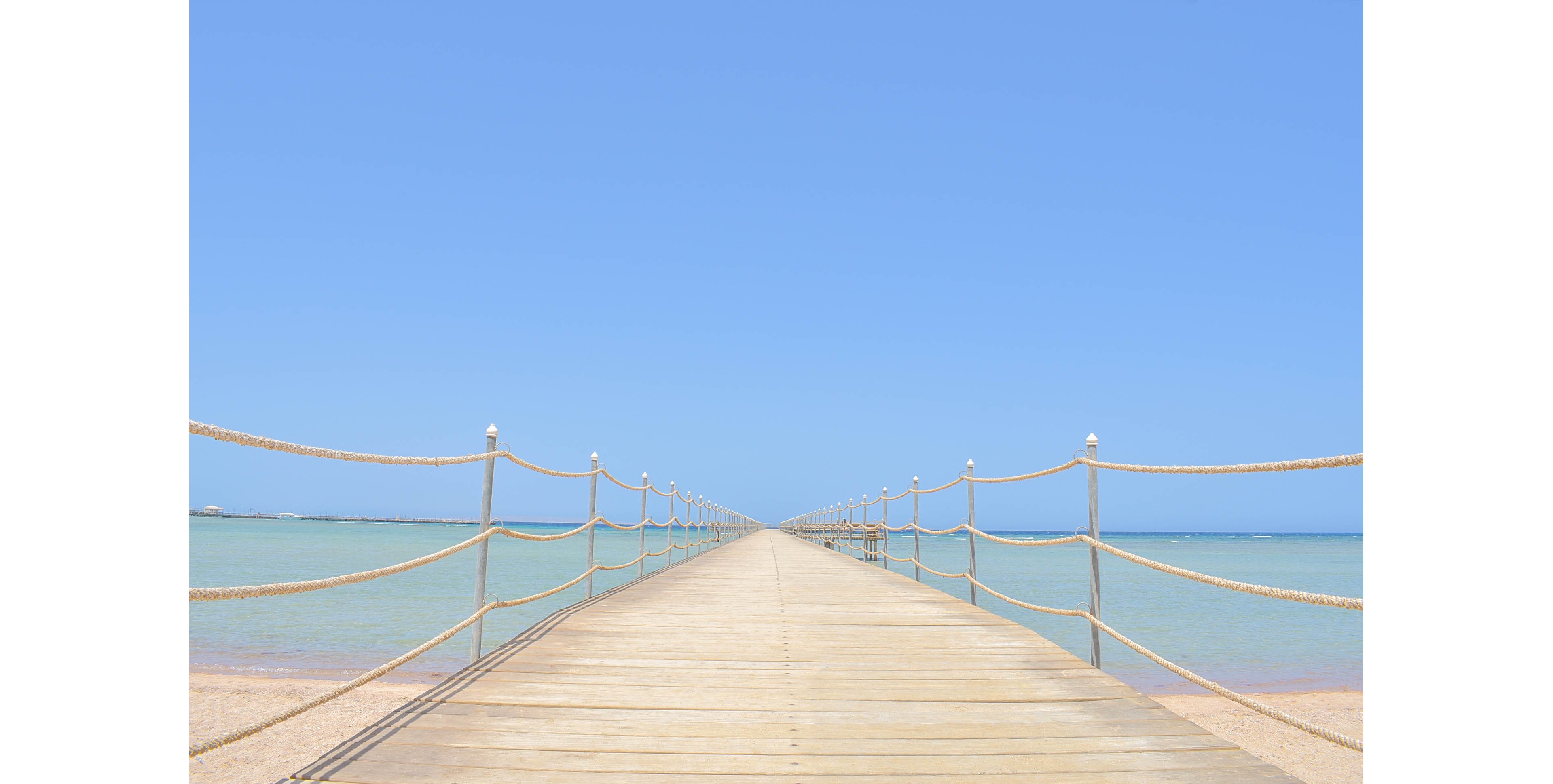 A long wooden pier stretches into the horizon over a calm, clear blue sea under a cloudless sky. The pier is bordered by simple rope railings, creating a serene and minimalist scene. The setting evokes a tranquil and summery coastal vibe.