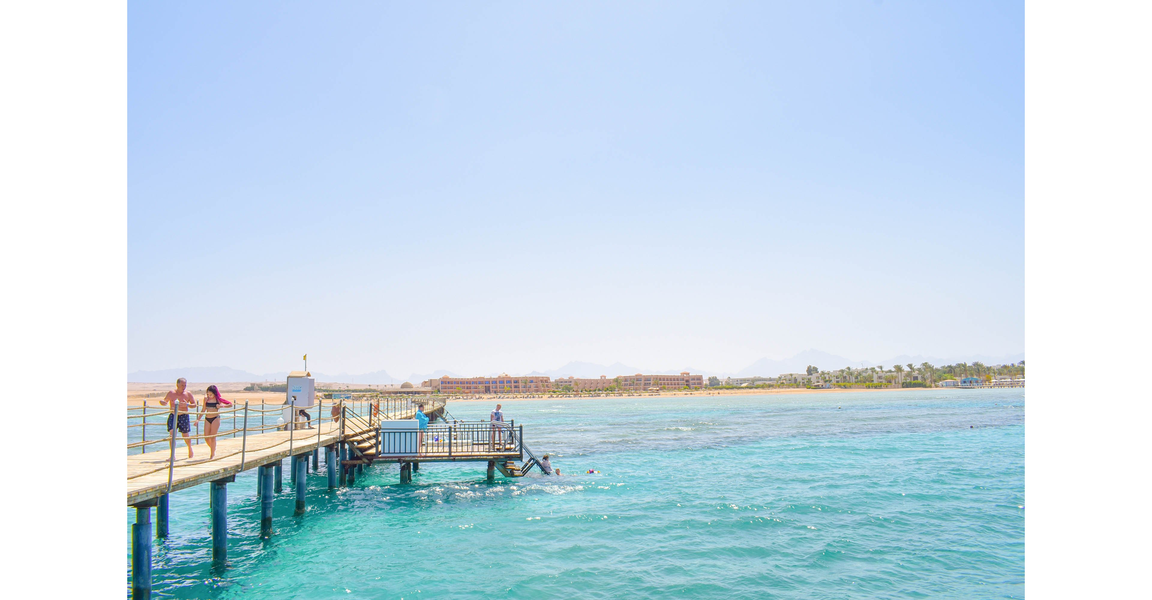 A scenic wooden pier extends over crystal-clear turquoise waters, with people enjoying the sunny day. The backdrop features a distant shoreline with buildings and palm trees under a bright blue sky. The serene setting suggests a relaxing coastal getaway.