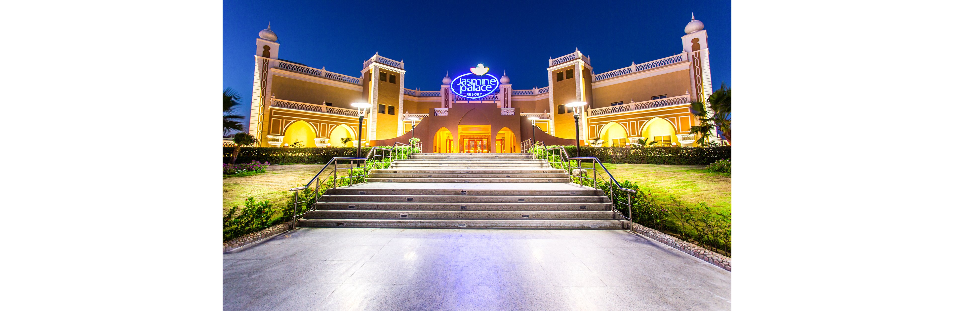 A grand architectural structure, Jasmine Palace, is brightly illuminated against a deep blue evening sky. The building features intricate design elements, arched entrances, and a prominent sign displaying its name. A wide staircase leads up to the entrance, surrounded by manicured greenery and decorative lighting.