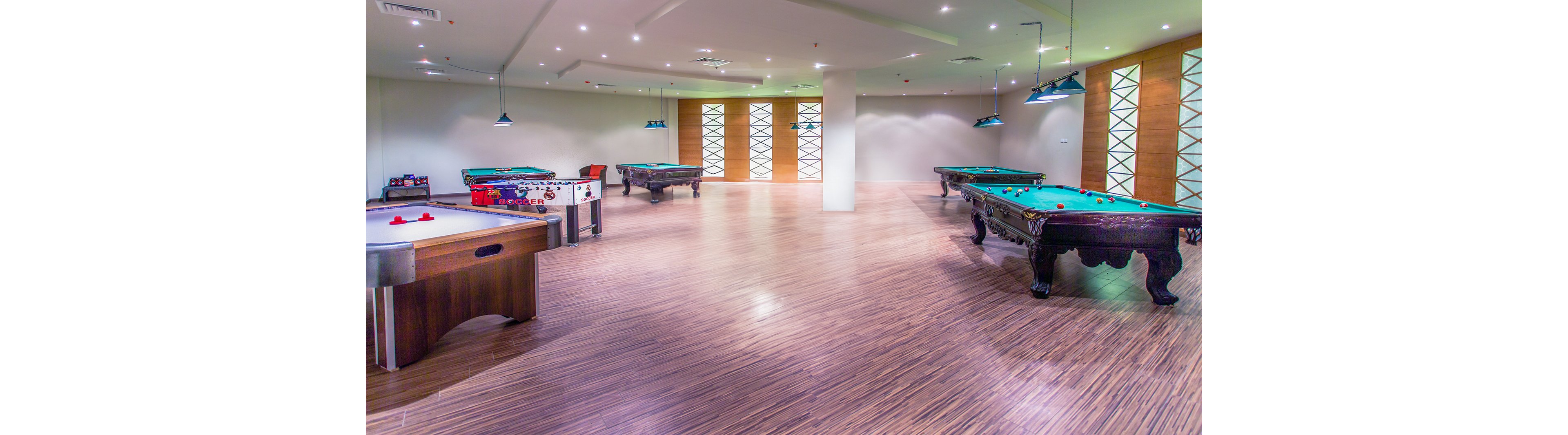 A spacious recreational lounge featuring multiple pool tables, air hockey, and foosball. A group of people, both men and women, are seated and standing, engaging in conversation and leisure activities. The room is well-lit with modern lighting and wooden flooring, creating a relaxed and inviting atmosphere.