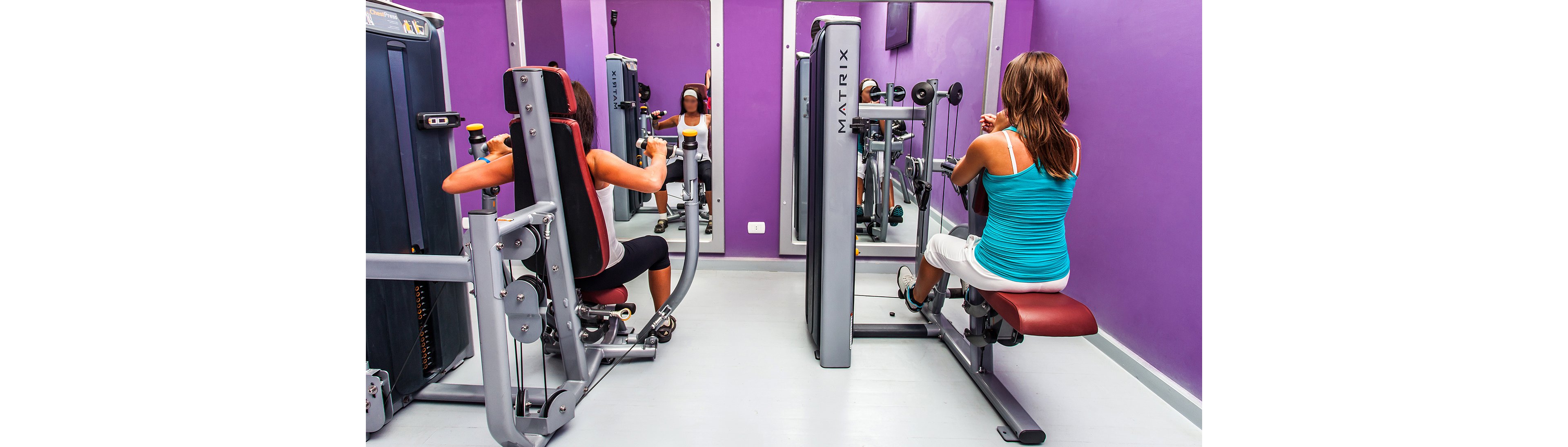 Two women are using strength training machines in a brightly lit gym with purple walls. The equipment includes a Matrix-branded machine, and the setting features mirrors and a clean, modern design. Both women are focused on their workout, creating an active and energetic atmosphere.