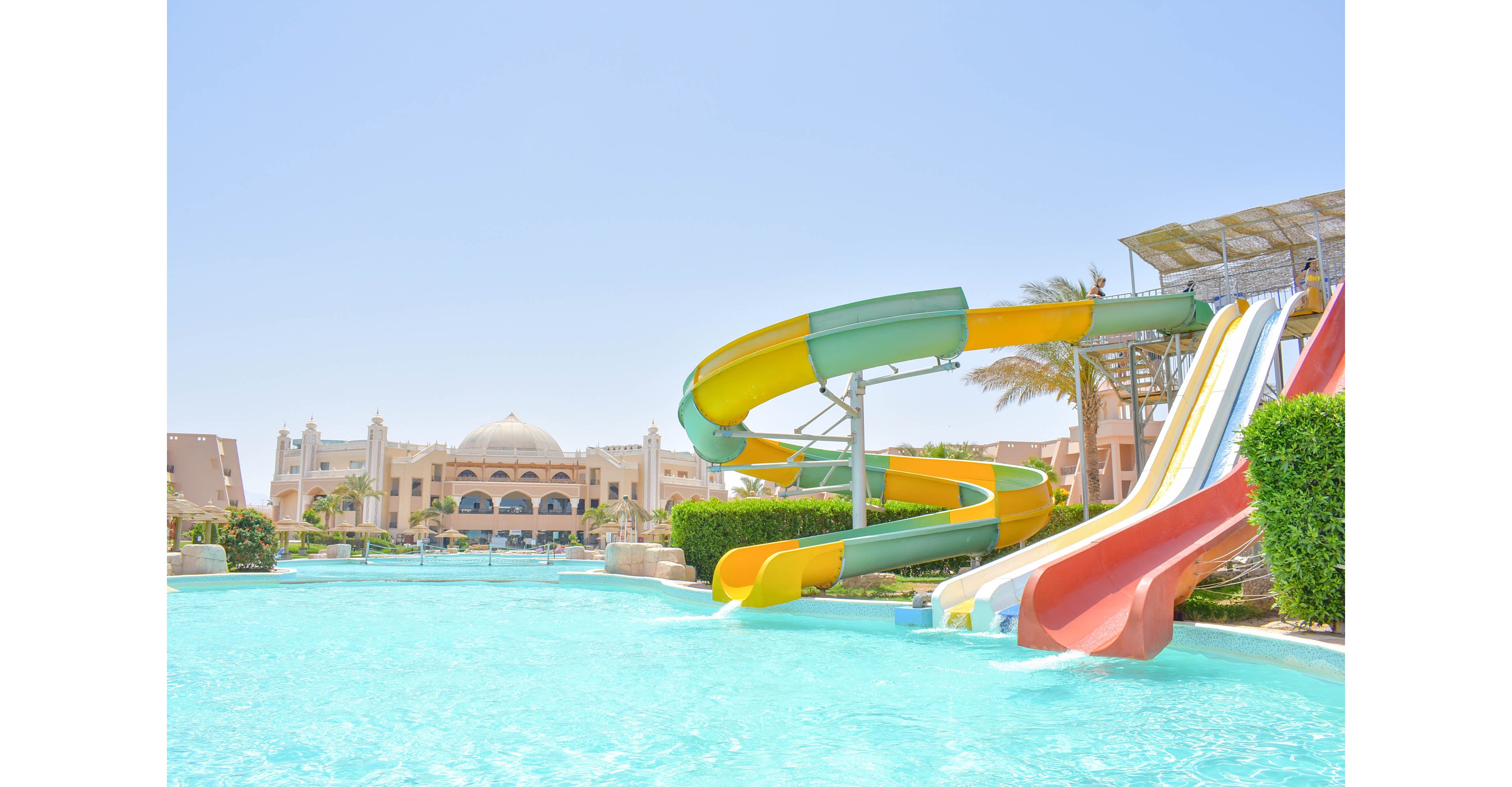 A vibrant water park featuring colorful slides leading into a clear blue pool, set against the backdrop of a luxurious resort with domed architecture. The sunny day enhances the bright and cheerful atmosphere. Palm trees and manicured greenery add to the tropical setting.