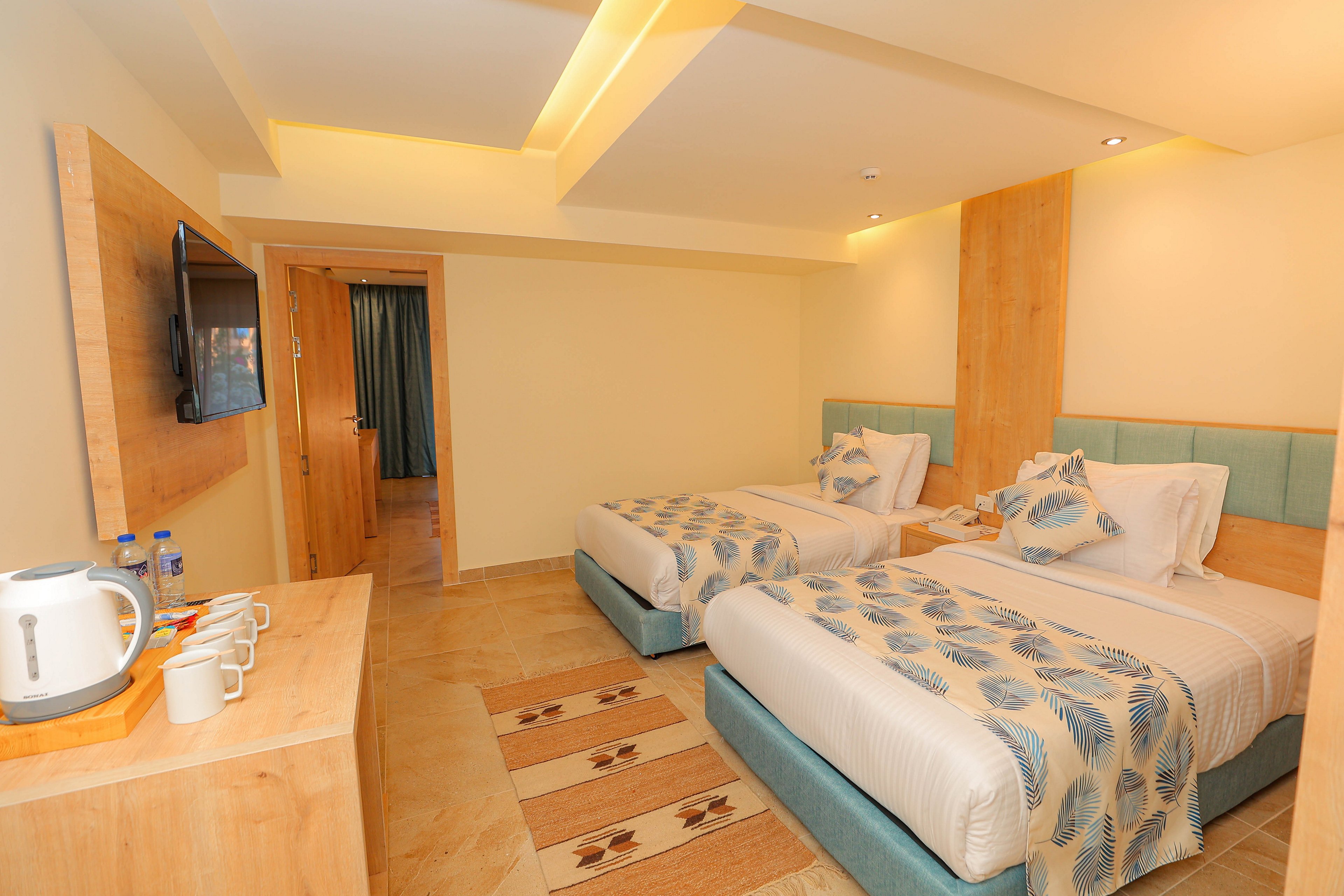 A cozy hotel room featuring two neatly made twin beds with patterned bedding. The room includes a wall-mounted television, a wooden desk with a kettle and cups, and soft ambient lighting. The decor is modern with light wood accents and a neutral color palette. A doorway leads to a curtain-covered area, adding depth to the space.