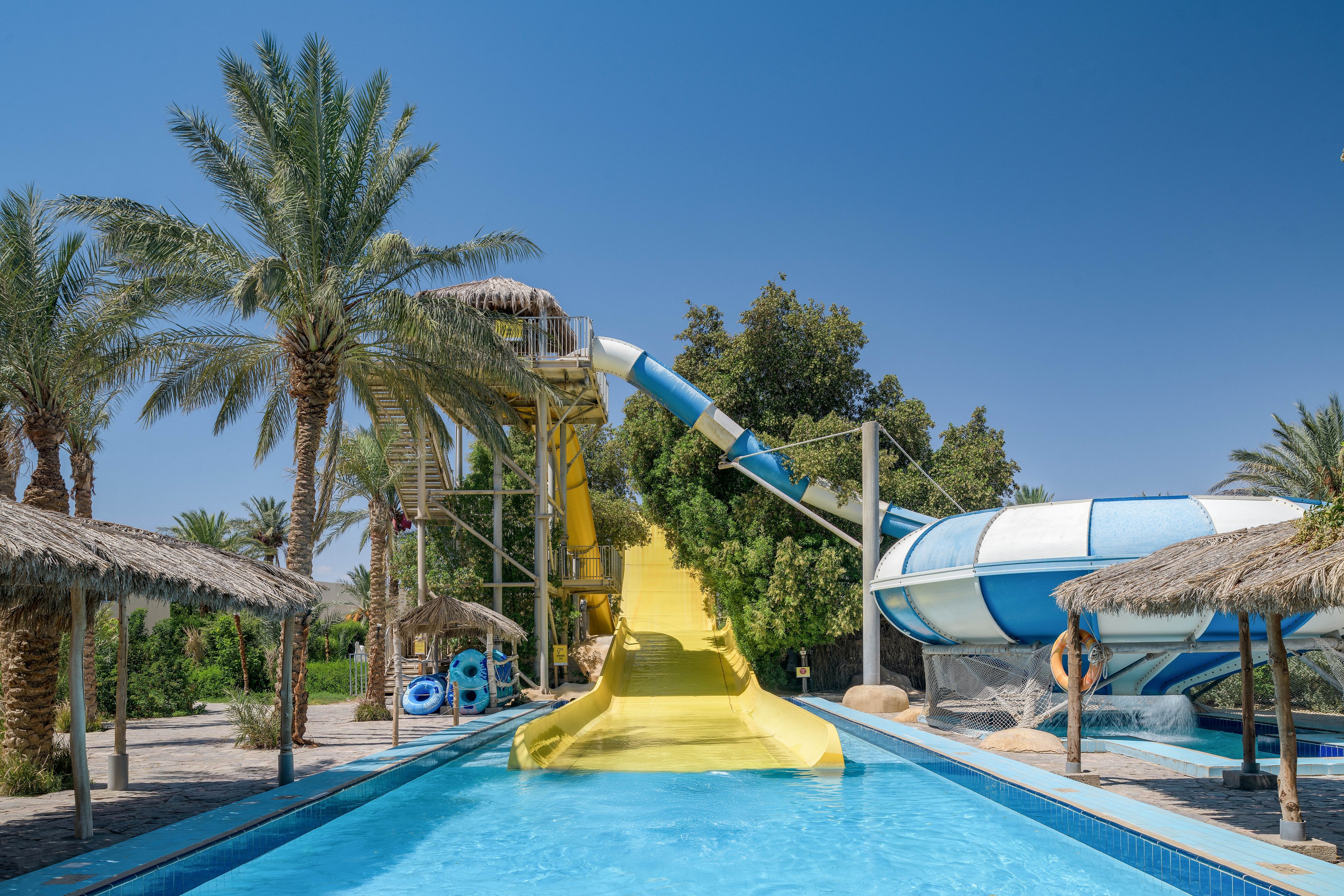 A family slides down a bright yellow water slide into a pool at a tropical resort. The setting features palm trees, thatched umbrellas, and a clear blue sky. A large spiral water slide is visible in the background, adding to the fun atmosphere.