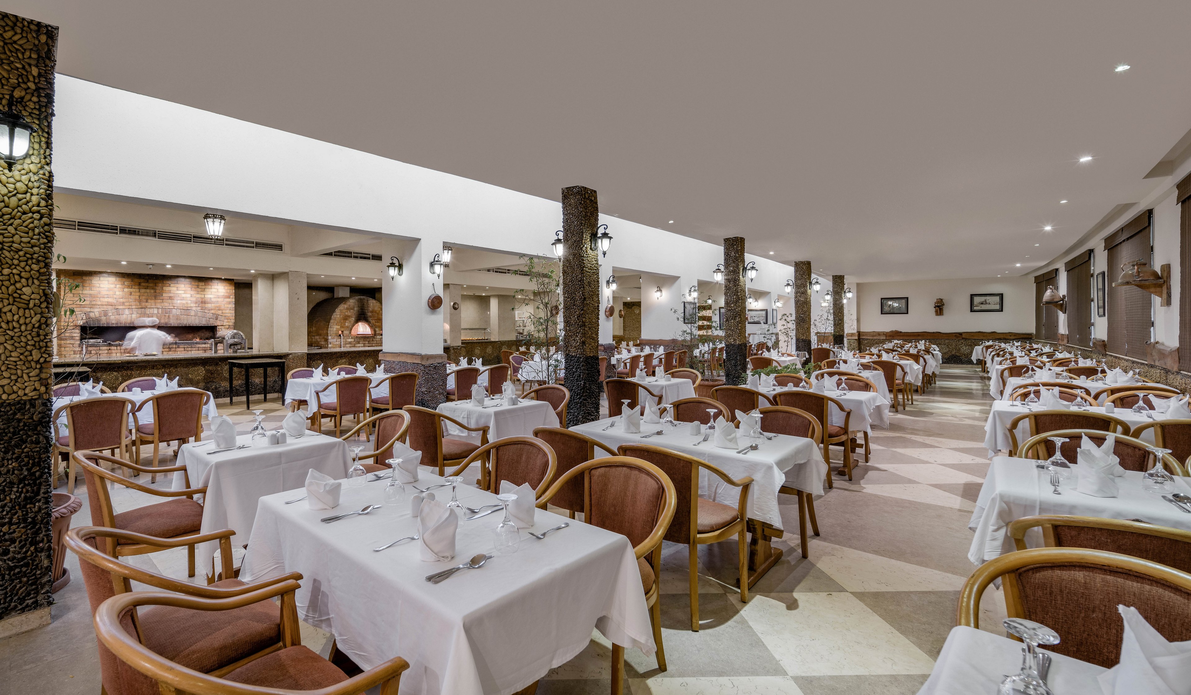 A well-lit and spacious restaurant dining area featuring neatly arranged tables with white tablecloths and wooden chairs. The setting includes a warm, neutral color palette and visible ceiling lights. The background showcases a buffet area with a rustic design, adding to the inviting ambiance.