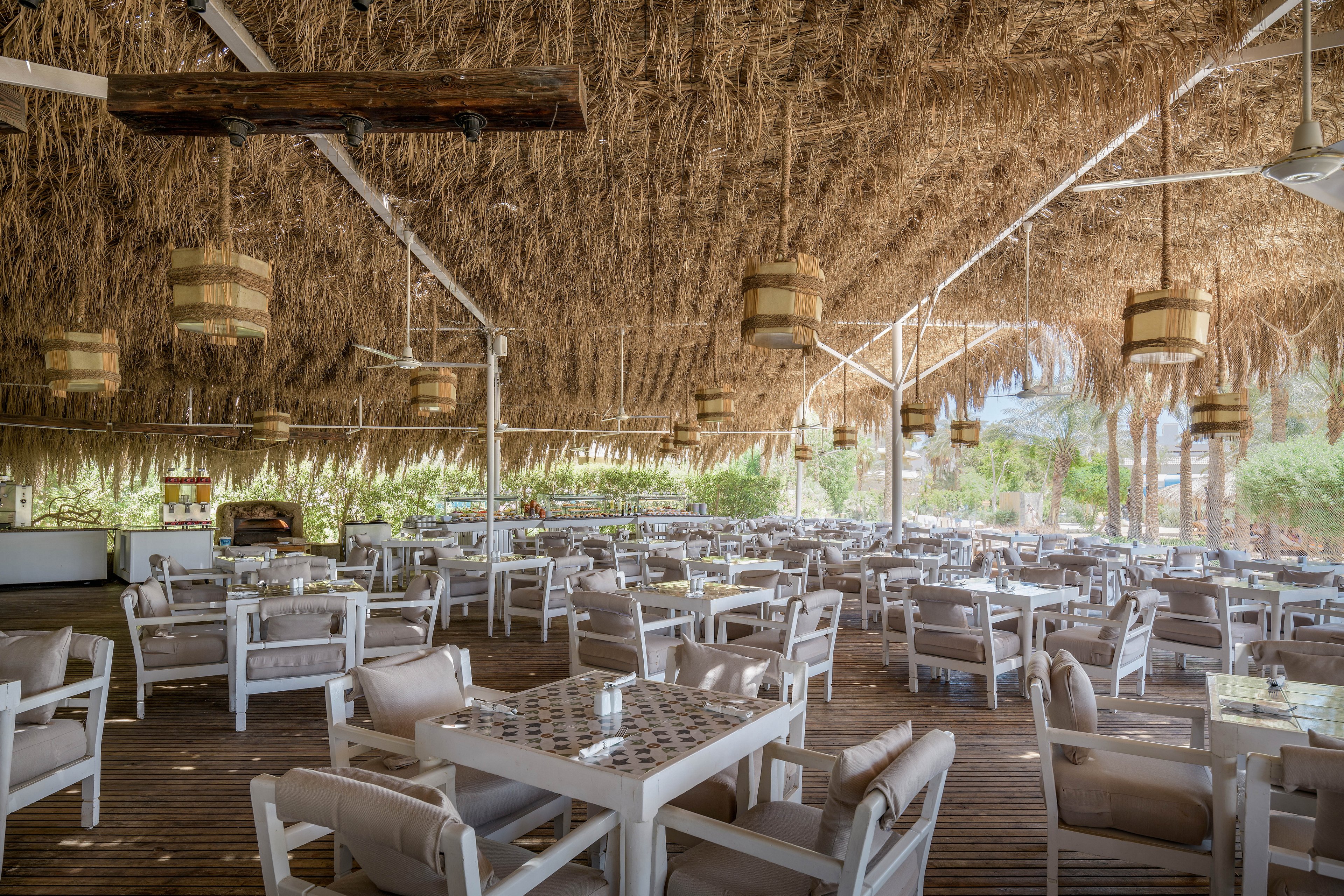 A spacious outdoor dining area featuring white wooden tables and chairs under a rustic straw-thatched roof. The setting is bright and airy, with natural light filtering through the open sides. Decorative hanging lights and lush greenery in the background enhance the relaxed and inviting atmosphere.