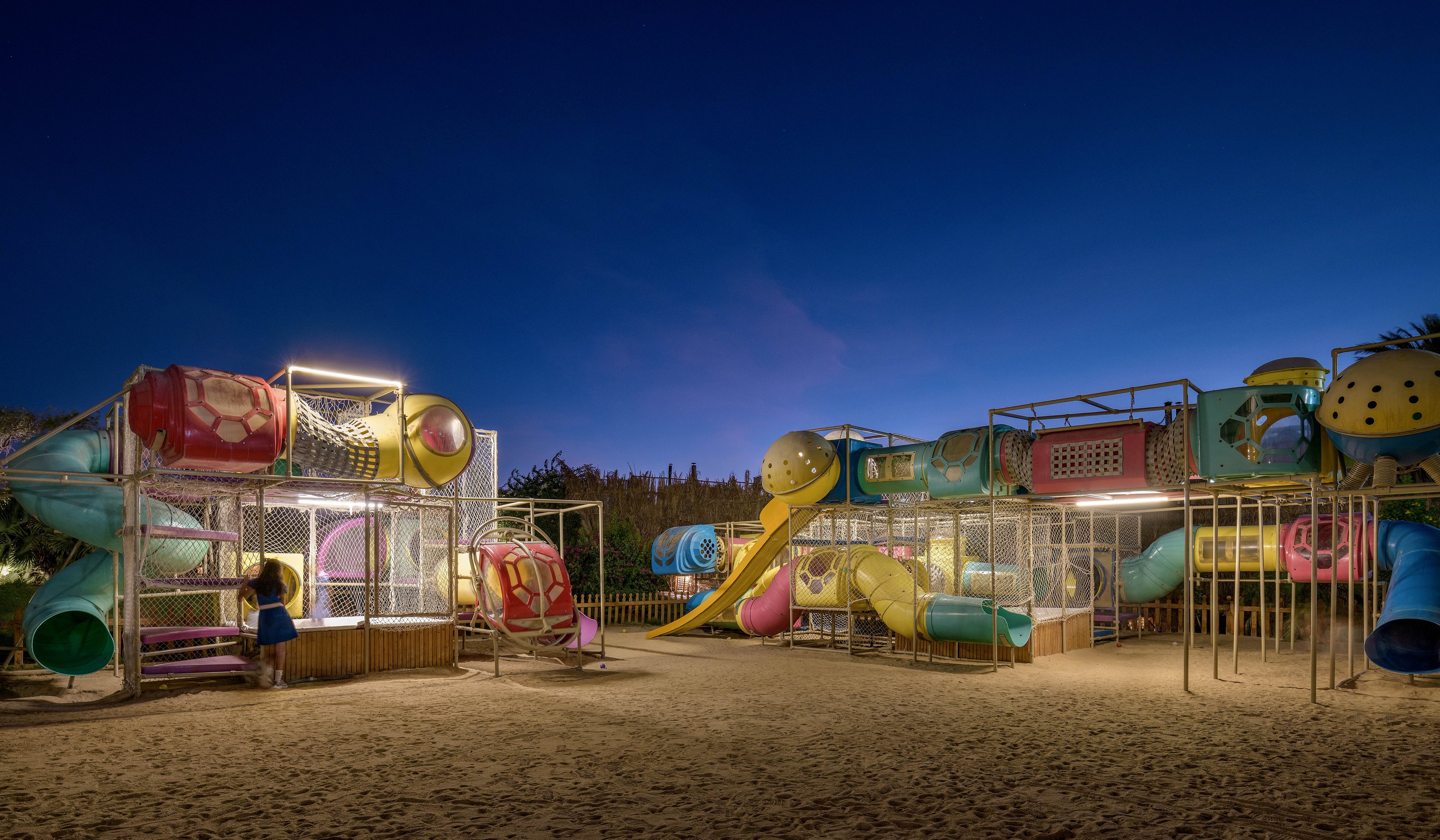 A brightly lit outdoor playground featuring colorful slides, tunnels, and climbing structures. The setting is a sandy area under a clear night sky, creating a serene and inviting atmosphere. The playground is empty except for one child exploring the structures.