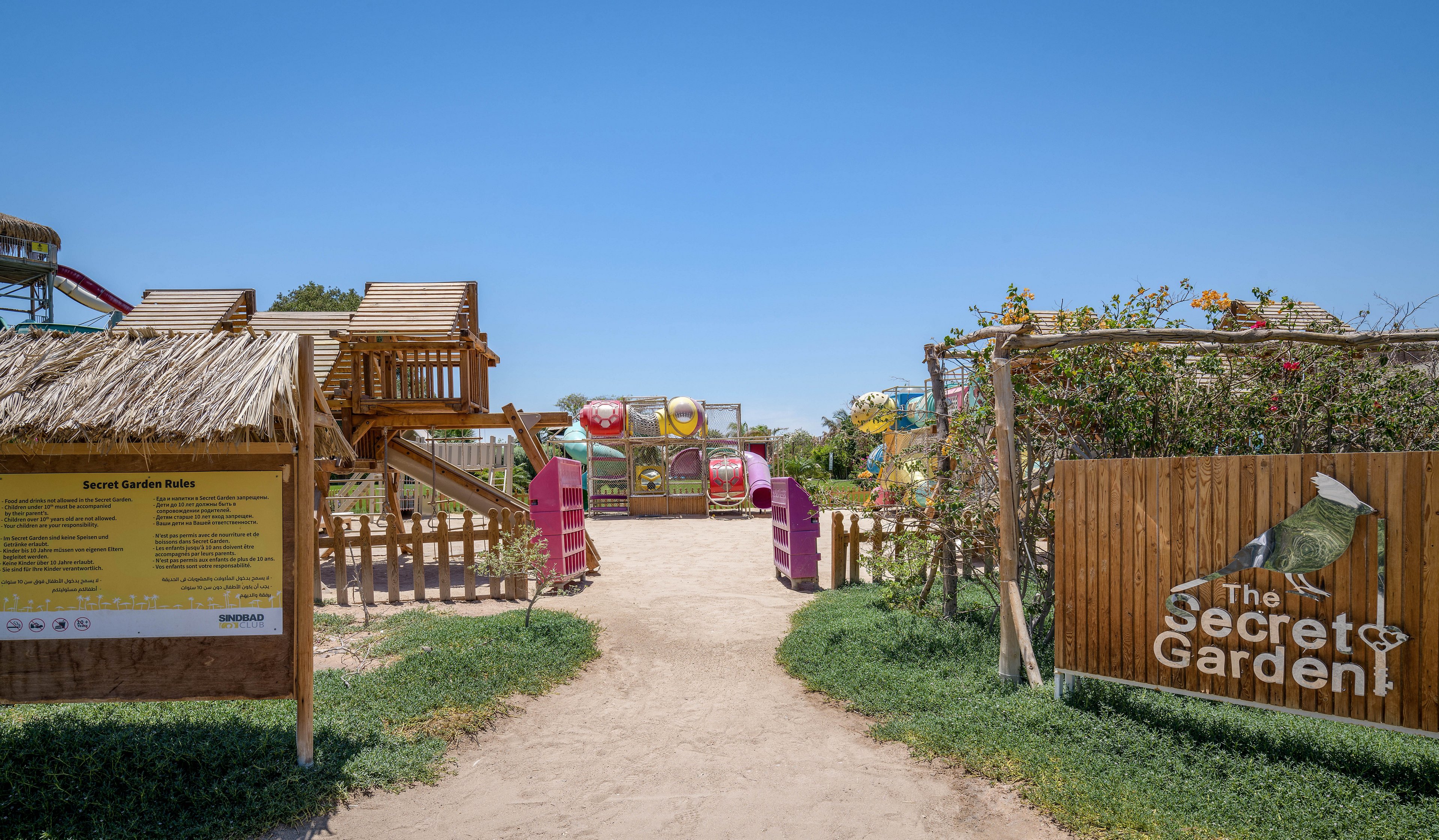 An inviting outdoor playground entrance featuring wooden play structures and a sign reading 'The Secret Garden.' The setting includes a clear blue sky, lush greenery, and colorful play equipment in the background. A large informational board is visible on the left side of the image.