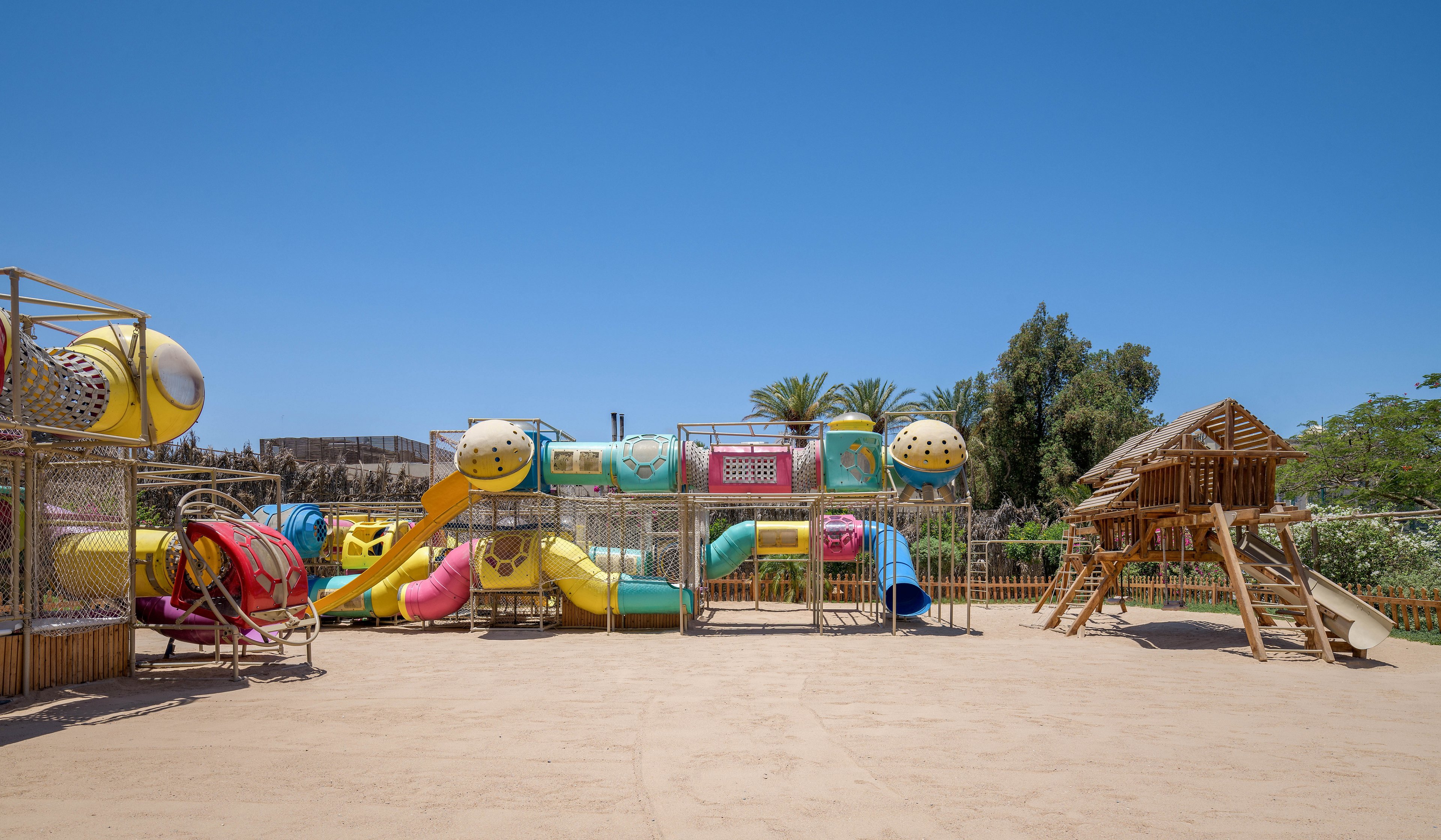 A vibrant outdoor playground featuring colorful slides, tunnels, and climbing structures under a clear blue sky. The setting includes a sandy ground and surrounding greenery, creating a fun and inviting atmosphere for children. The wooden playhouse adds a rustic touch to the modern play equipment.