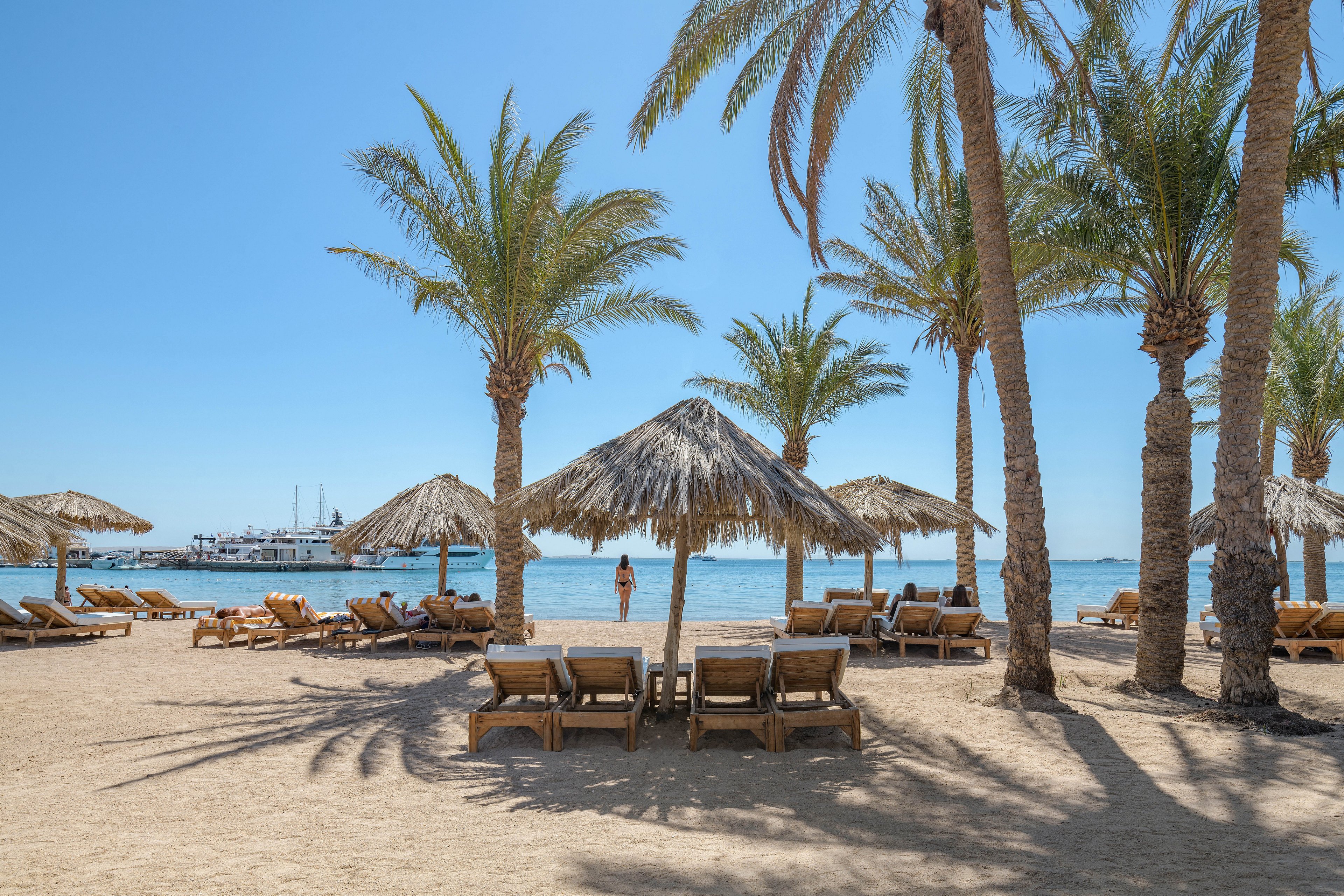 A serene tropical beach featuring palm trees, thatched umbrellas, and wooden loungers arranged on the sandy shore. A person stands near the water, with a boat visible in the distance under a clear blue sky. The setting evokes relaxation and leisure in a sunny coastal environment.