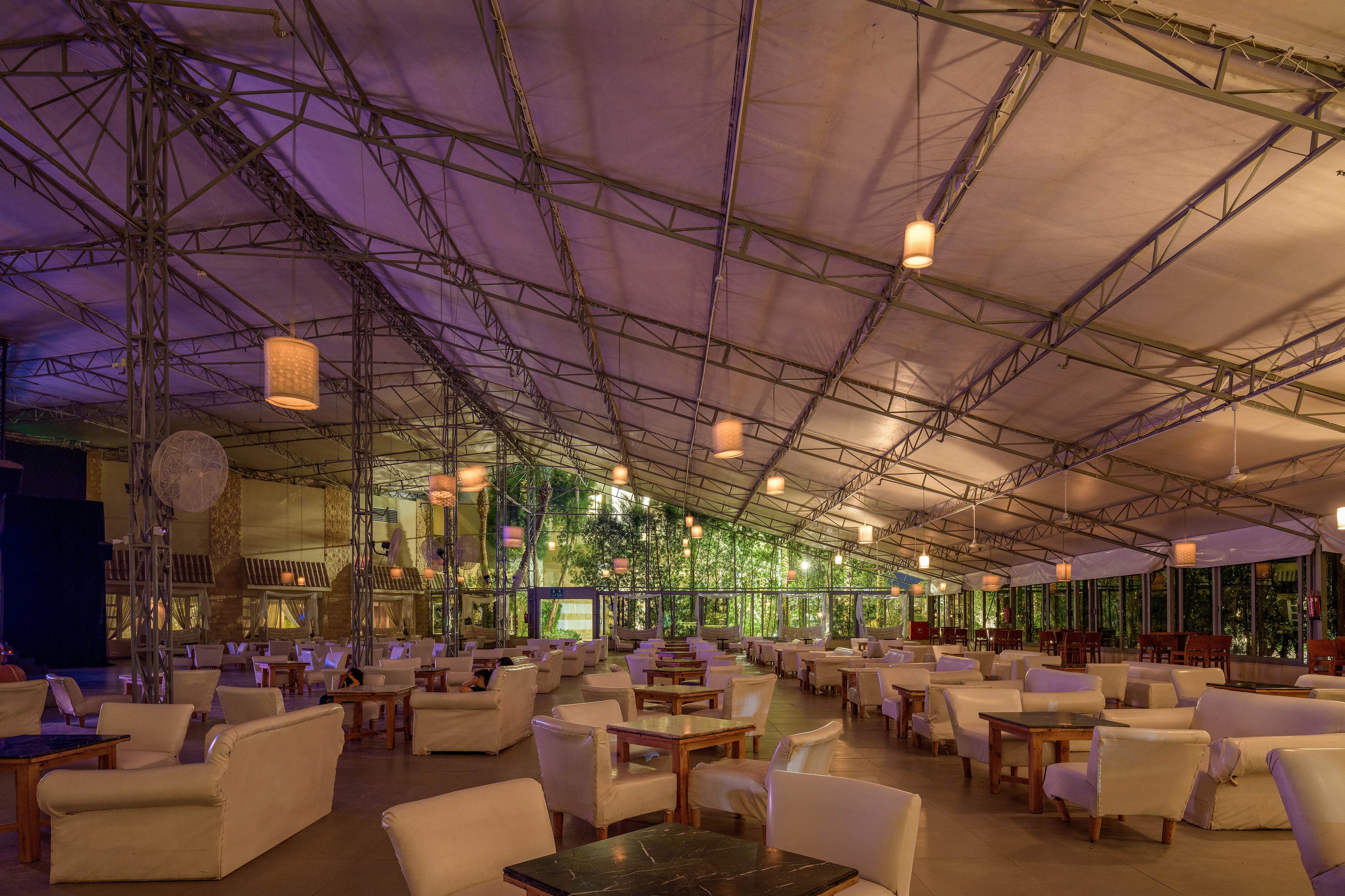 A large, open-air lounge area with a high canopy roof and soft ambient lighting. The space features comfortable white armchairs and wooden tables arranged in a relaxed layout. The setting is illuminated by hanging pendant lights, creating a warm and inviting atmosphere. Greenery is visible in the background, enhancing the natural and tranquil vibe.