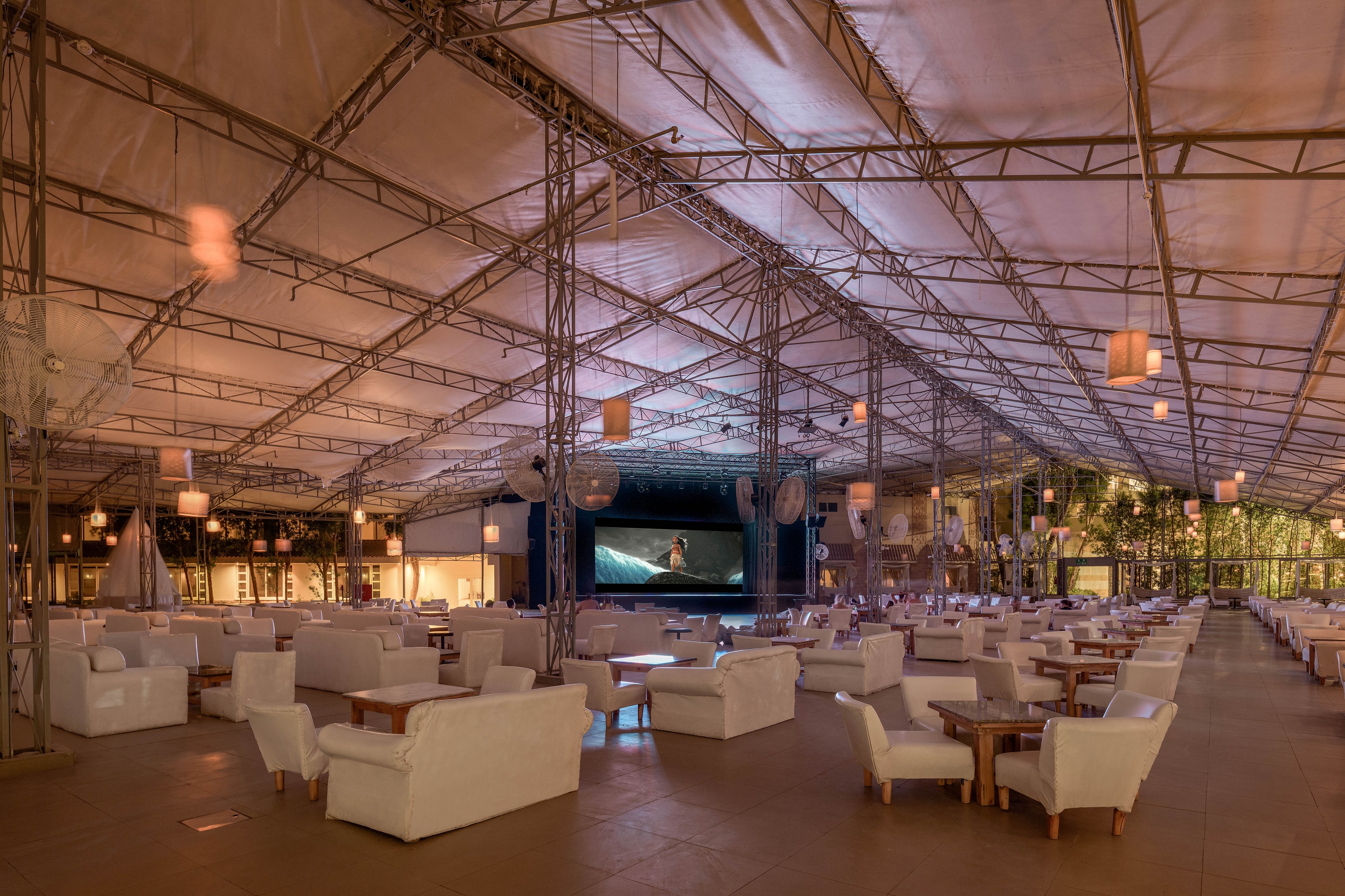 A spacious outdoor lounge area featuring white sofas and wooden tables under a large canopy. A large screen is visible at the far end, displaying a movie scene. The setting is illuminated with warm hanging lights, creating a cozy and inviting atmosphere.