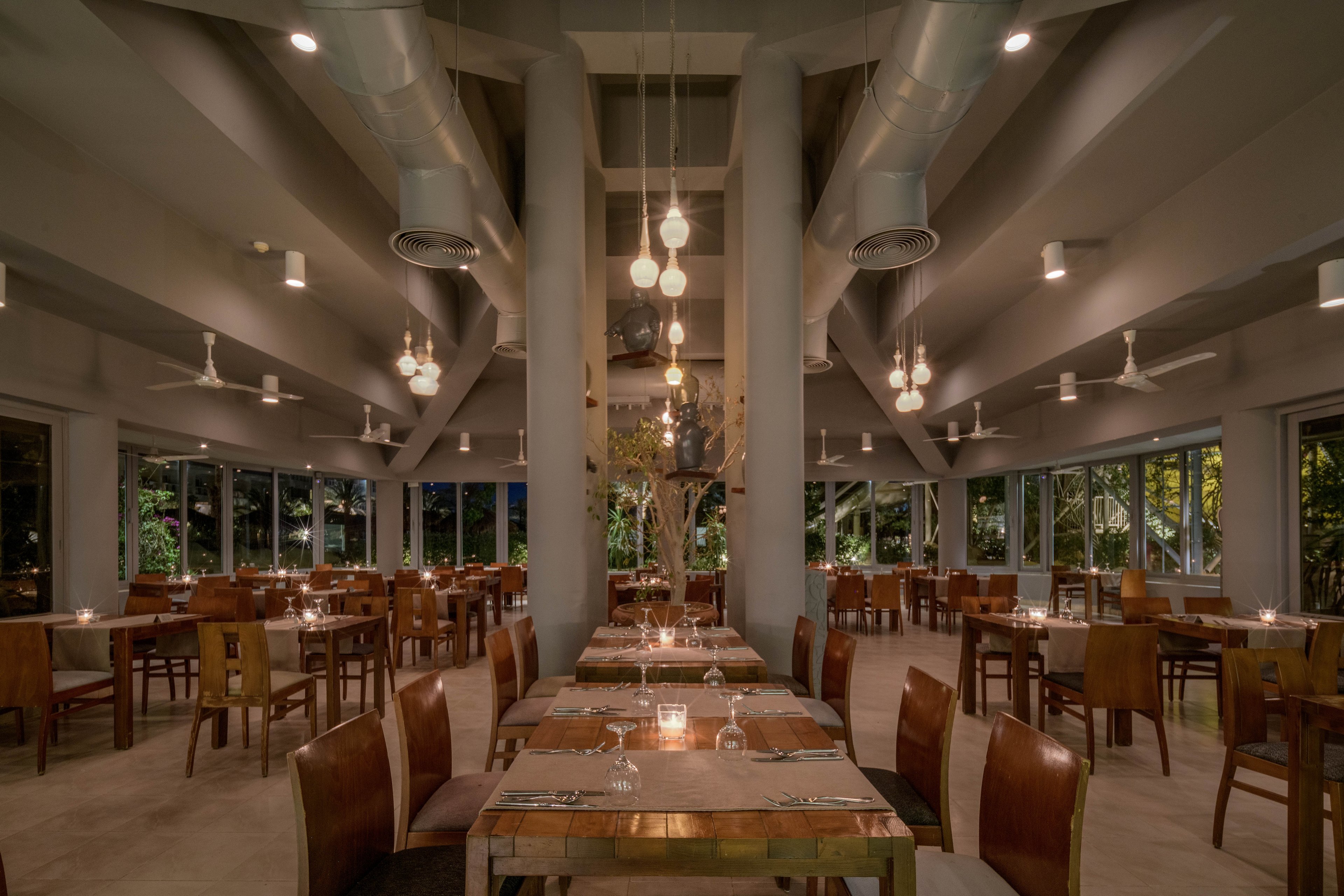 A spacious restaurant interior featuring wooden tables and chairs arranged neatly under warm pendant lighting. The setting includes large windows offering views of greenery outside, creating a cozy and inviting atmosphere. The ceiling showcases exposed ducts and fans, adding a modern industrial touch.