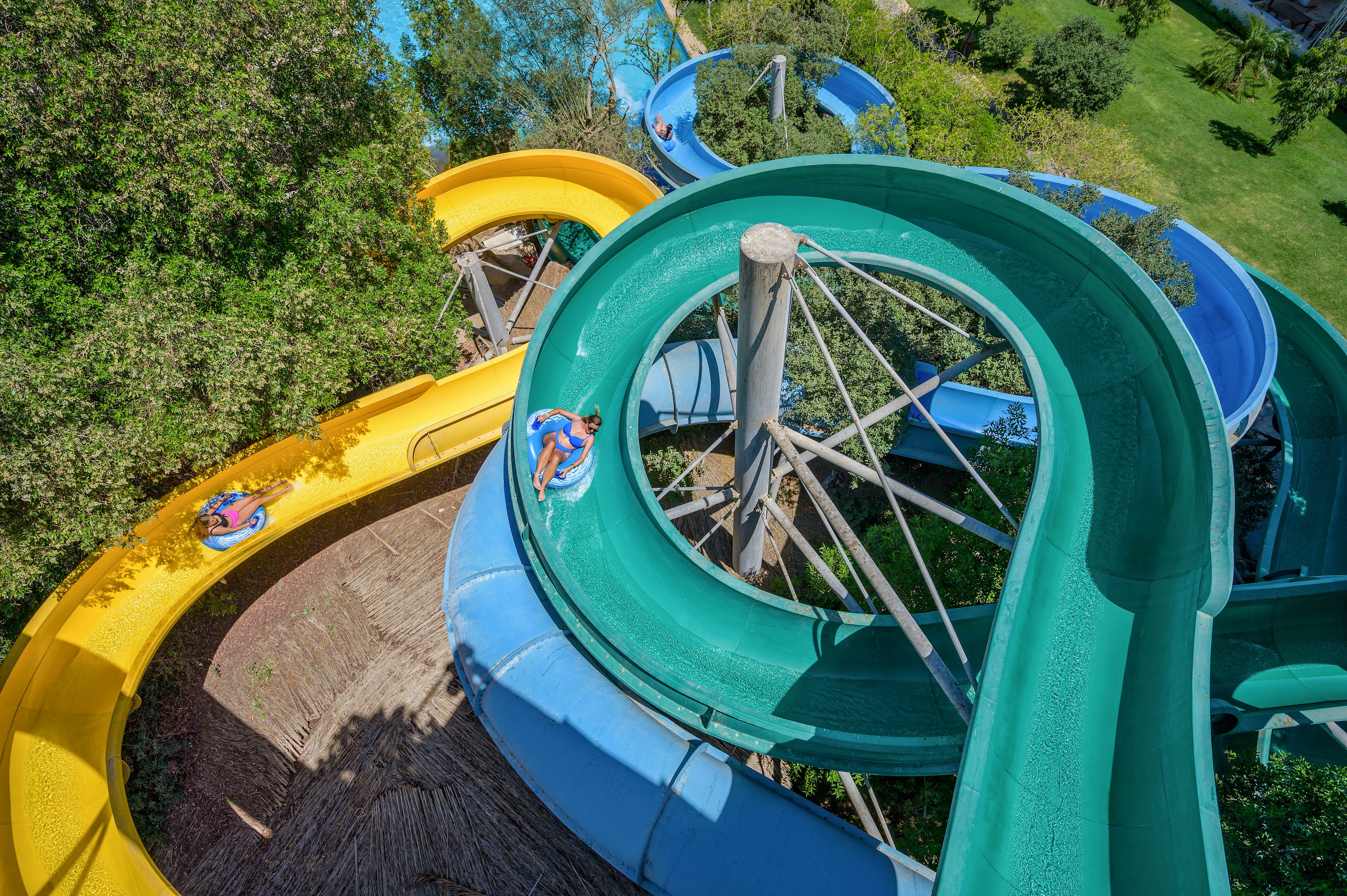 A vibrant outdoor water park featuring intertwined yellow, green, and blue slides surrounded by lush greenery. The slides curve and twist, creating an exciting visual of dynamic movement. The setting is sunny and inviting, ideal for recreational activities and family fun.