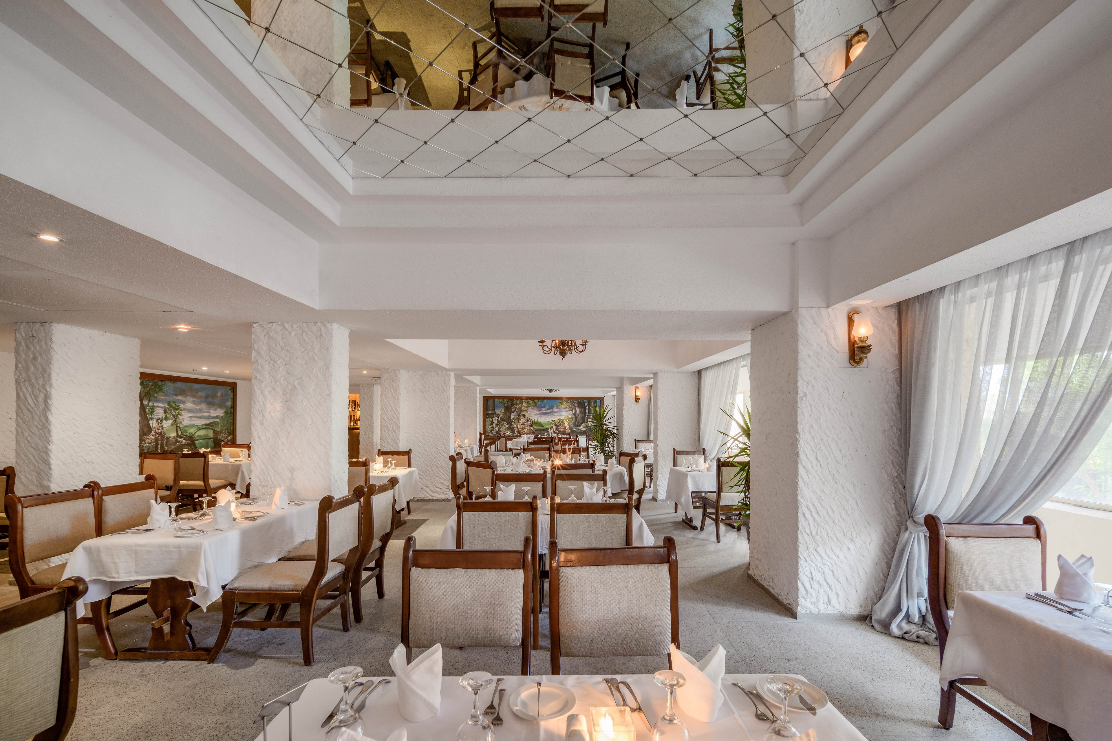 A spacious and elegant restaurant interior featuring white walls, tablecloths, and classic wooden chairs. The setting includes large windows with sheer curtains, allowing natural light to brighten the space. The upper level is visible with additional seating, and decorative paintings adorn the walls, adding a touch of sophistication.