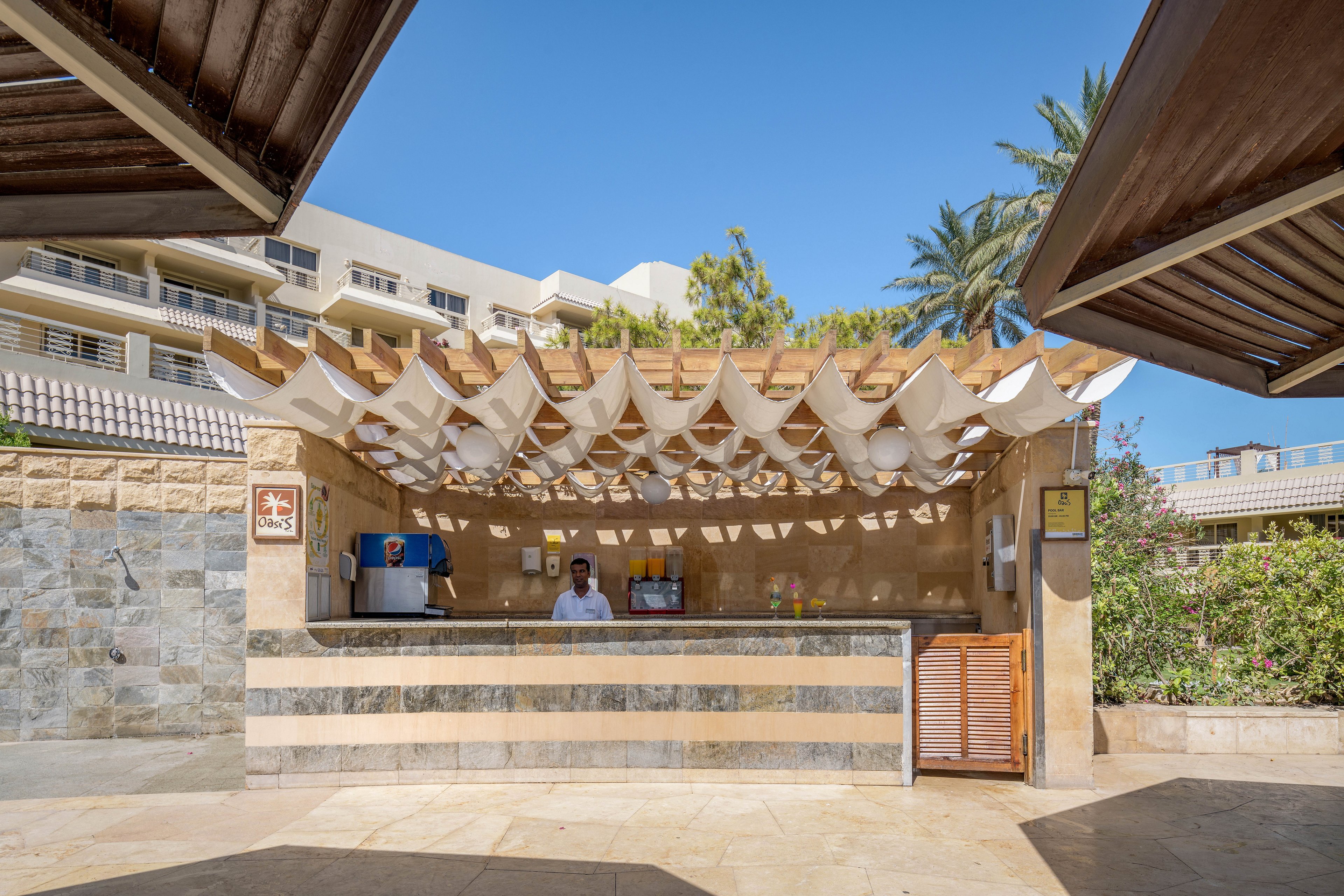 An outdoor bar with a wooden canopy featuring decorative fabric panels, located in a sunny resort setting. A bartender is standing behind the counter, ready to serve. The background includes resort buildings, lush greenery, and a clear blue sky, creating a relaxed and inviting atmosphere.